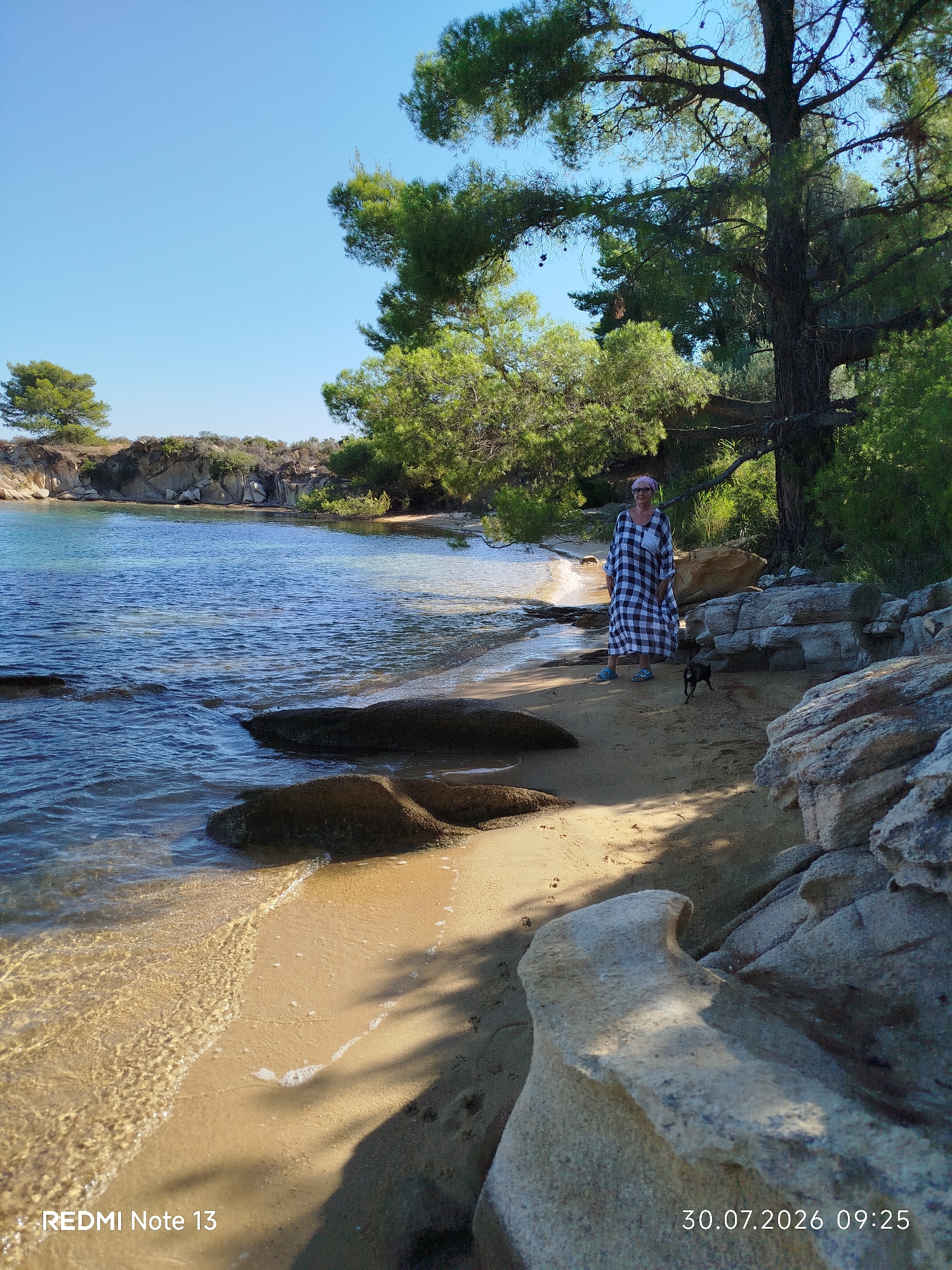 A woman and a small black dog are on a sandy beach near Sithonia Municipal Unit, Greece. The woman is wearing a black and white checkered dress and a pink headscarf. She is standing near the water's edge, and the dog is in front of her. The beach is lined with rocks and trees. The water is clear and blue. The sky is clear and blue. The image was taken on July 30, 2026, at 9:25 AM.