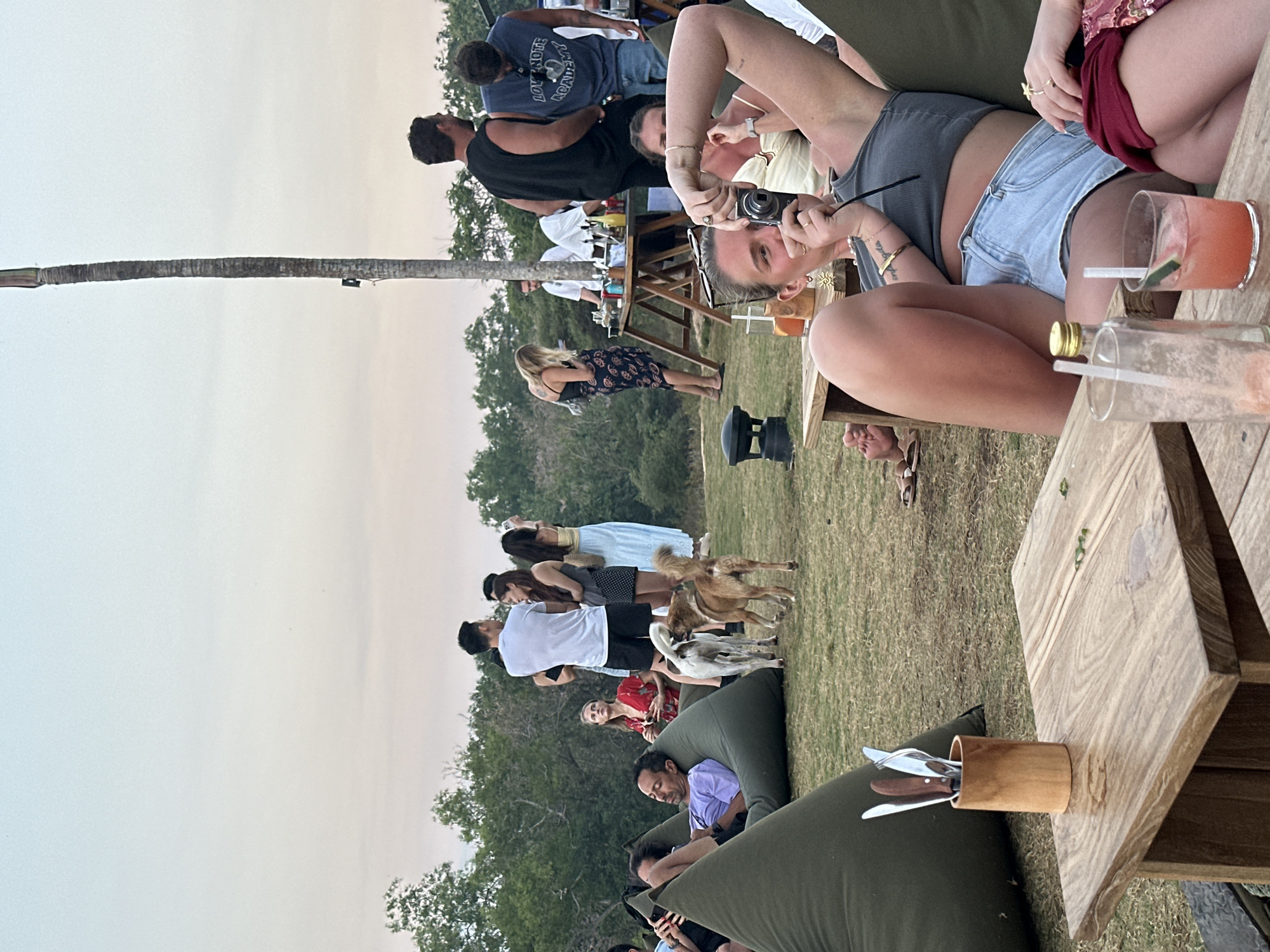 In Kuta Selatan, Indonesia, at dusk, a group of people are gathered on a grassy knoll, enjoying a social event with drinks and food. Several individuals are seated on large, olive-green beanbag chairs, while others stand and converse. A wooden table in the foreground holds drinks and cutlery. In the background, a bar area is set up with bottles and containers, and a tall palm tree stands against the light sky. Two dogs are present in the scene, interacting with the people. The overall atmosphere appears relaxed and festive.