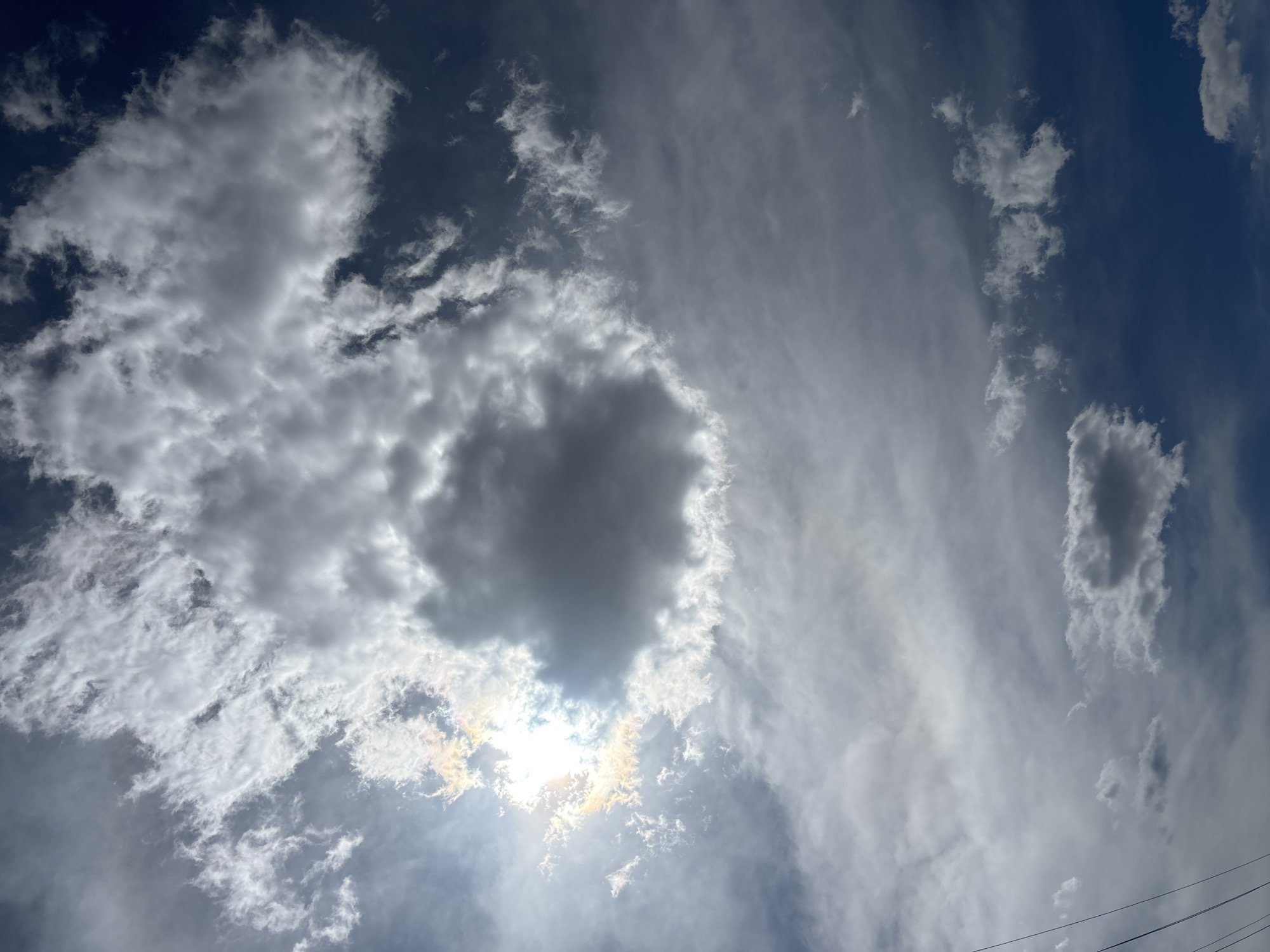 The image shows a view of the sky, filled with a variety of cloud formations. Predominantly, the clouds are grey and white, with a large, dark cloud partially obscuring the sun. The light from the sun is filtered and diffused, creating a shimmering effect. Some areas of the sky are a clear, bright blue. The image was taken in Zagatuy, Russia. There are powerlines shown in the bottom right of the frame.