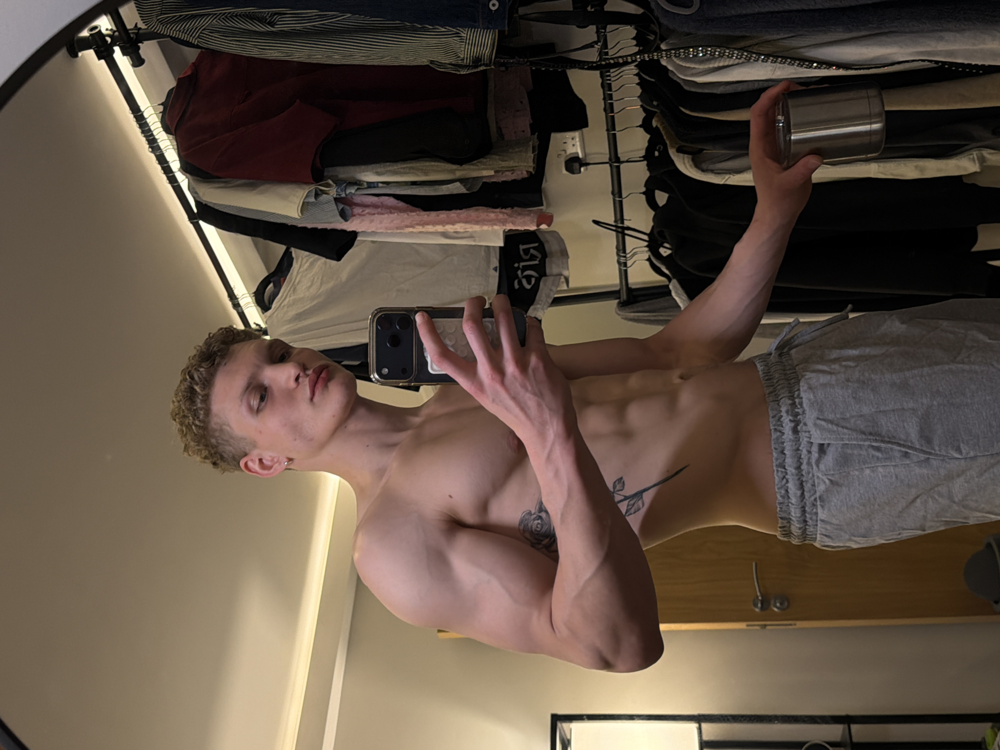 A shirtless male with fair skin, short blonde curly hair, and tattoos on his left arm and abdomen is taking a mirror selfie in a room. He is wearing grey sweatpants. In the background, a closet with various clothes hanging is visible, along with a door and some other furniture. The location cannot be determined to be London, United Kingdom from the context of the image.