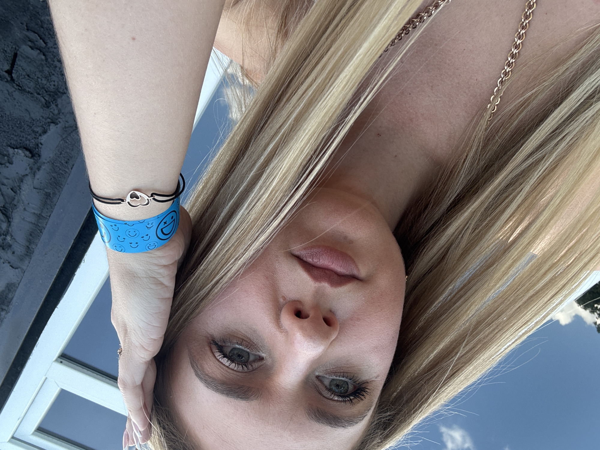 A blonde woman is taking a selfie outdoors. She is wearing a blue wristband with smiley faces and a silver heart charm bracelet. Her hand is on her forehead. The background shows a window and the sky.