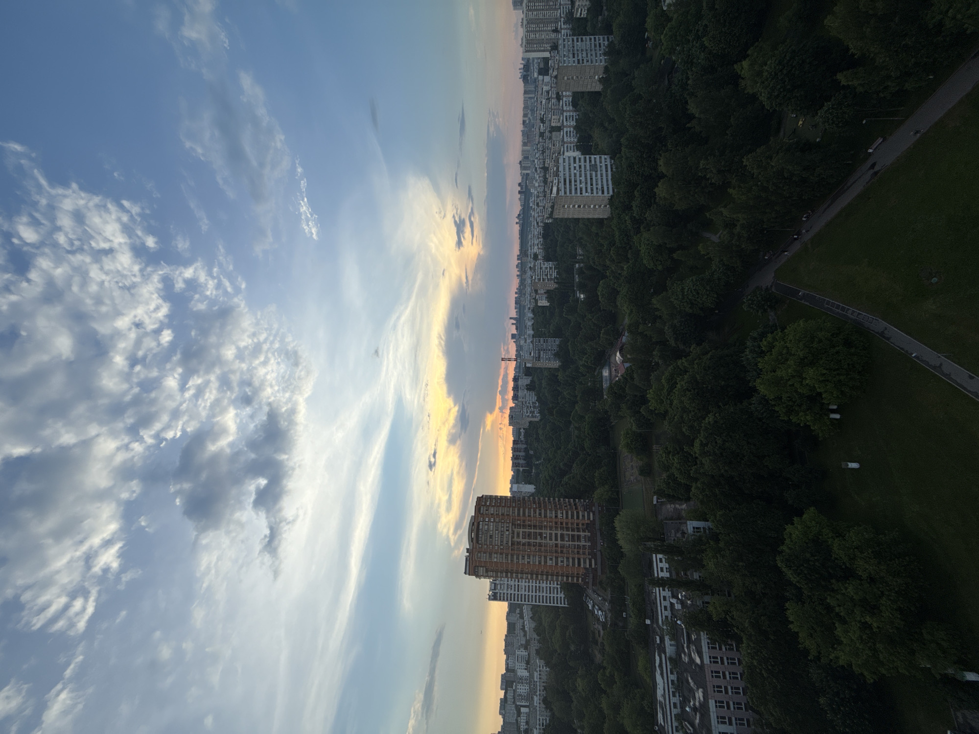 The image is an aerial view of a city, likely Moscow, Russia, during a sunset or sunrise. The sky is a vibrant mix of blues, whites, and oranges, with scattered clouds. In the foreground, there are dense green trees and a park area with pathways. Several residential buildings, ranging in height and architectural style, are visible throughout the cityscape. A prominent tall, orange-brick building stands out among the others. The overall scene depicts a typical urban environment with a significant amount of green space, suggesting a well-planned residential area. There are no discernible people or specific actions captured in the frame.