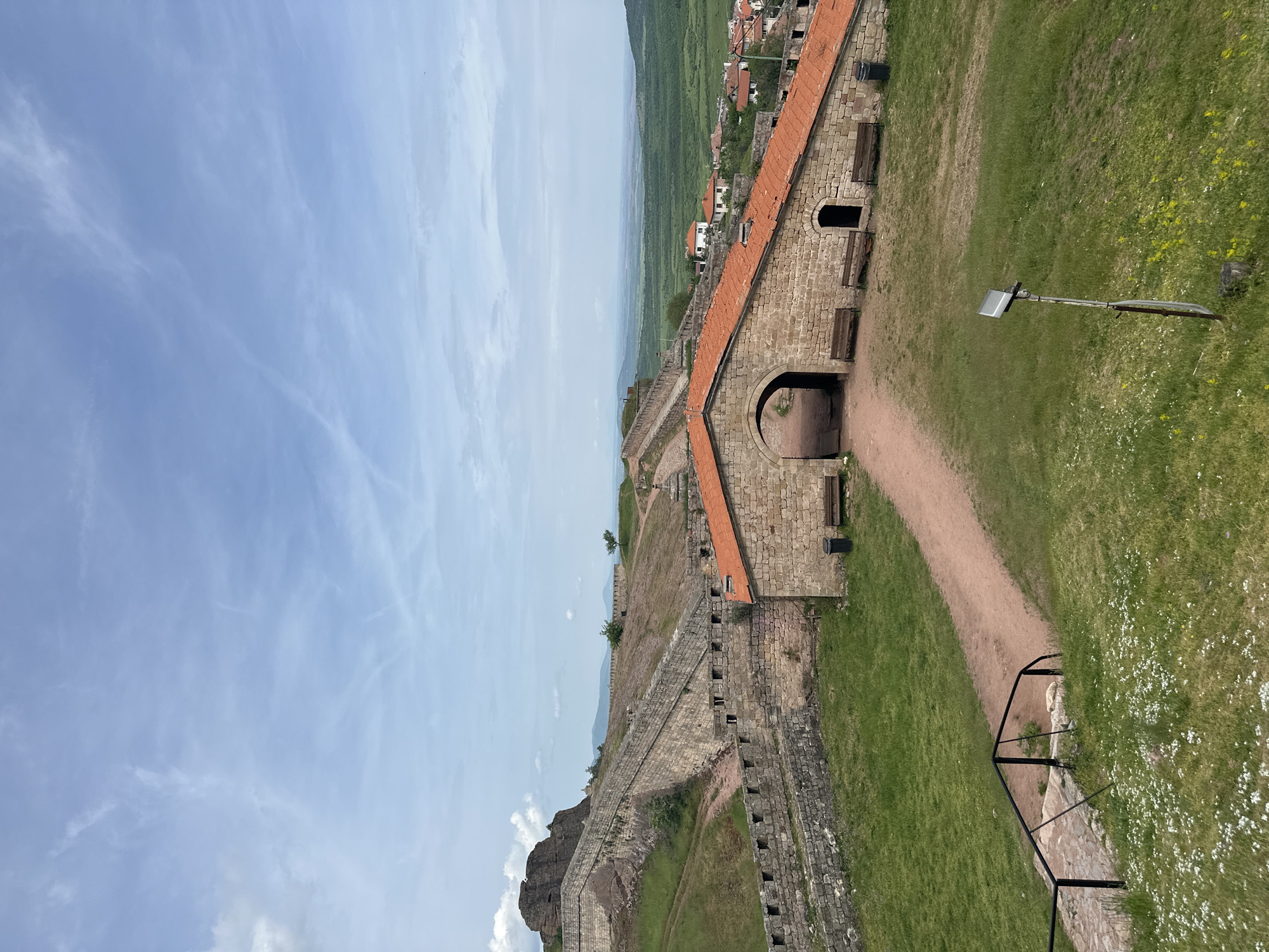 A high-angle shot displays a portion of the Belogradchik Fortress in Belogradchik, Bulgaria. The image depicts a historical brick and stone structure with a red-tiled roof stretching alongside a grass-covered area. Part of the fortress wall is visible, extending in the distance towards a town and a landscape featuring rolling green hills. A path is seen along the fortress leading to an arched entrance. The sky is partially cloudy and light blue.