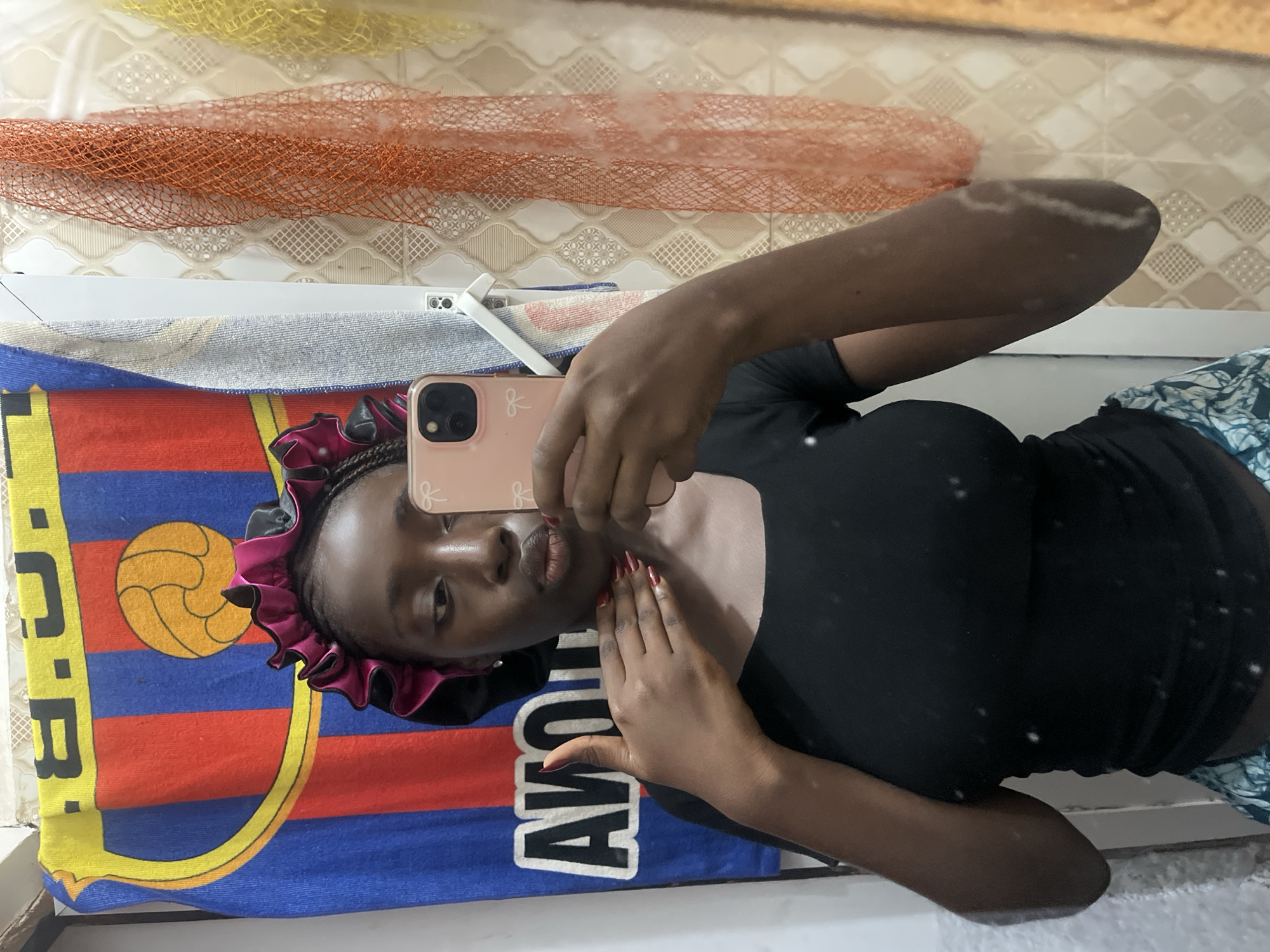 The image shows a person taking a selfie in what appears to be a bathroom. The person has dark skin, a pink hair bonnet, and is wearing a black top. They are holding a pink phone to take the picture. In the background, there is a patterned tile wall, a towel with the FC Barcelona logo, an orange netting material, and a white towel rack. It is not possible to confirm the location as Abuja, Nigeria with only the provided image.