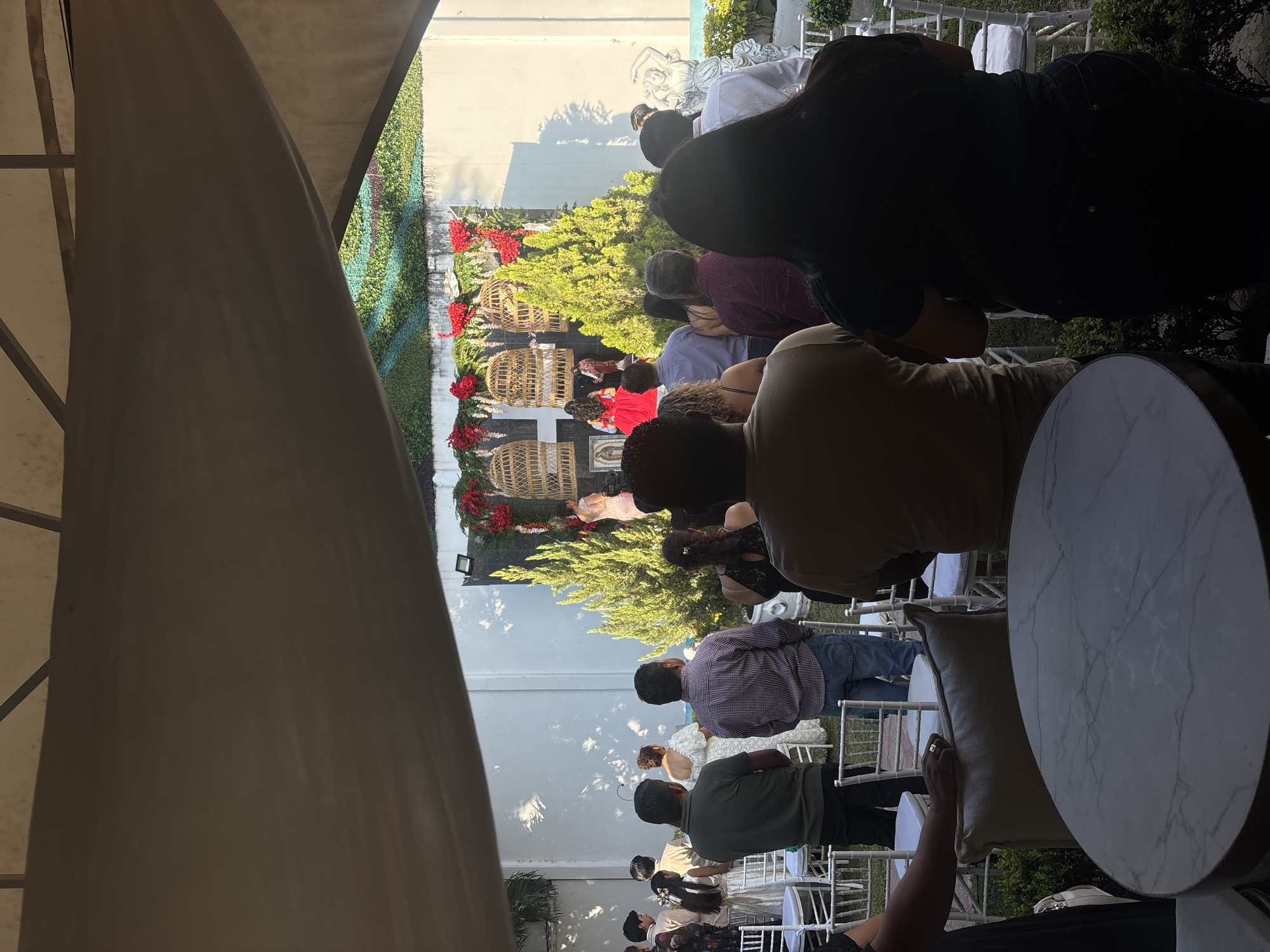 The photograph depicts an outdoor event, possibly a wedding or ceremony, in Chilpancingo, Mexico. Numerous people are seated in rows of white chairs, facing towards a decorative arrangement of greenery, wicker cages, and red floral accents. The attendees are diverse in attire. A white canopy partially obscures the view from the left. A round marble-topped table is visible in the foreground, suggesting a reception or social gathering. The scene takes place in an outdoor setting, possibly a garden or courtyard. The location appears to be near a wall, possibly a building or property border.