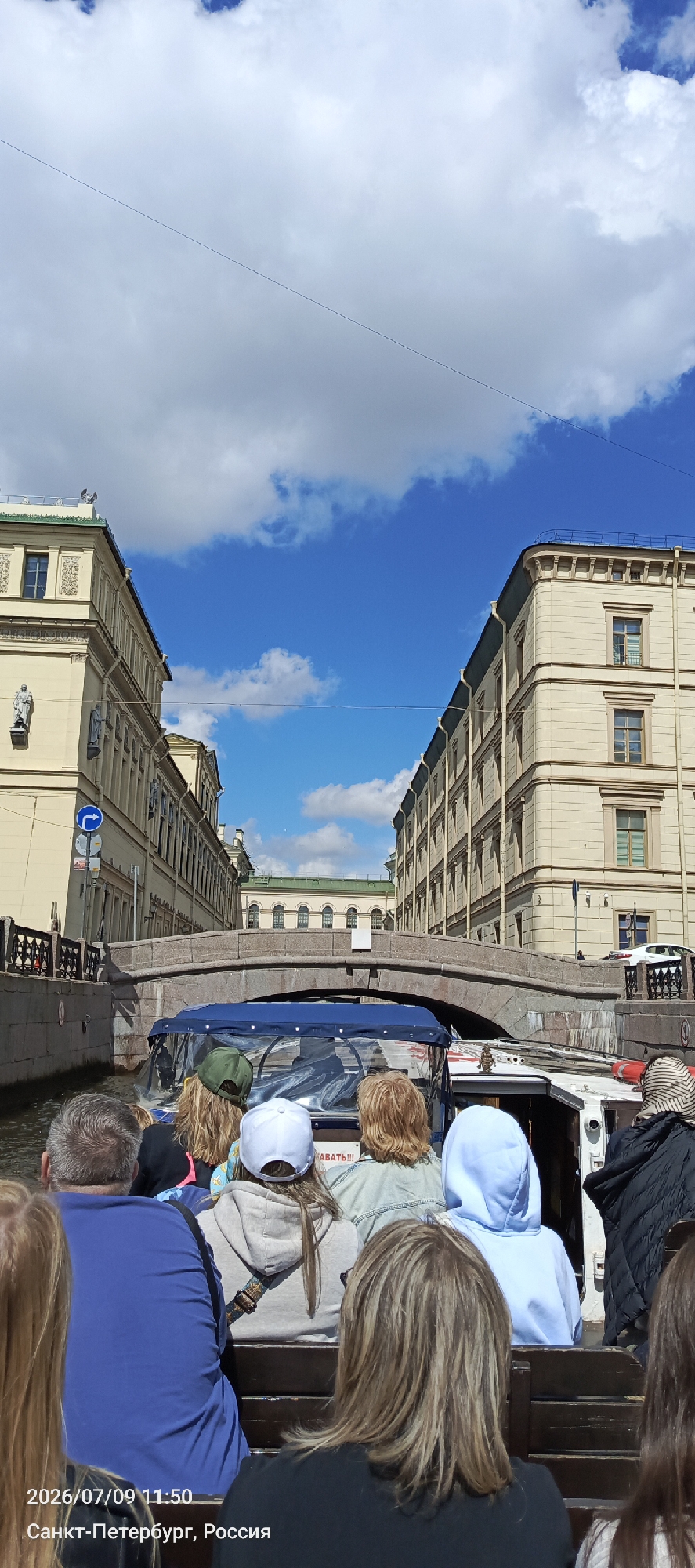 The photograph captures a perspective from a boat traveling on a canal in Saint Petersburg, Russia. The boat is filled with passengers facing away from the camera, with their backs to the viewer. The canal is bordered by historic buildings with ornate architecture, typical of Saint Petersburg. A stone bridge arches over the canal, connecting the two sides of the street. The sky is bright blue with scattered clouds, indicating a pleasant day. The timestamp and location "2026/07/09 11:50 Санкт-Петербург, Россия" are overlaid on the lower left portion of the image.