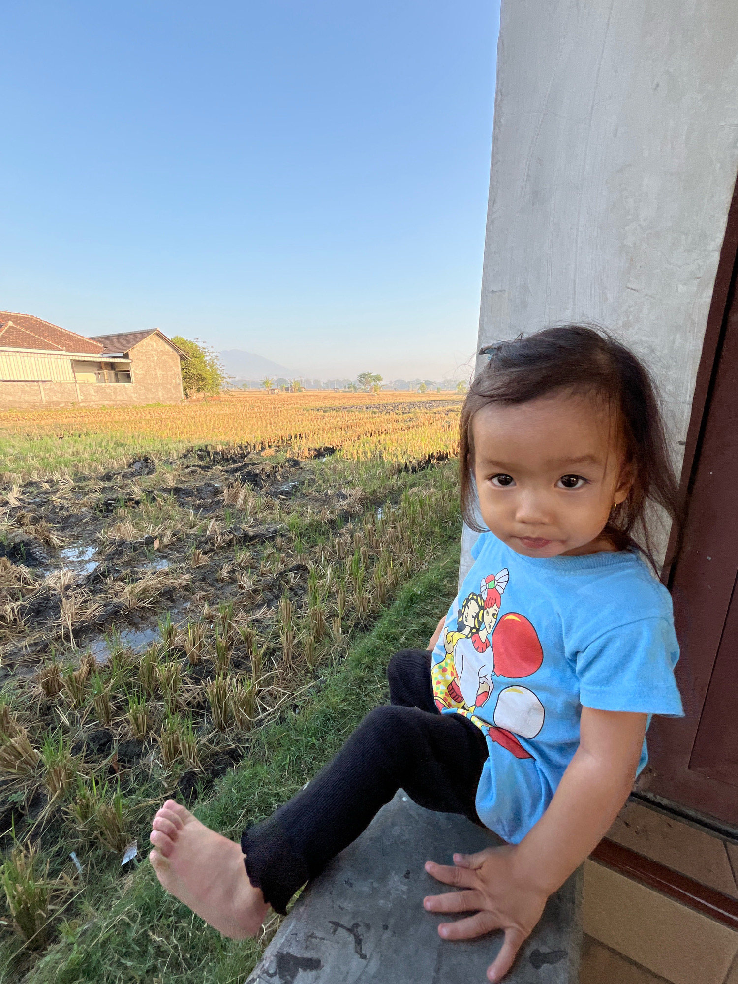 In the city of Kauman, Indonesia, a young girl with dark hair and brown eyes sits on a concrete ledge, facing slightly towards the viewer. She is wearing a bright blue t-shirt with a cartoon graphic and black leggings. Her left foot is bare and extended forward, revealing her toes. Behind her, a rural landscape unfolds. Stubble from a harvested rice field stretches into the distance, interspersed with muddy patches and some standing water. Two houses with tiled roofs are visible in the mid-ground, with a hazy mountain range in the far background under a clear blue sky. The girl is positioned on the right side of the frame, leaning against a light-colored wall and a brown doorway. Her right hand rests on the concrete ledge in front of her, fingers spread.