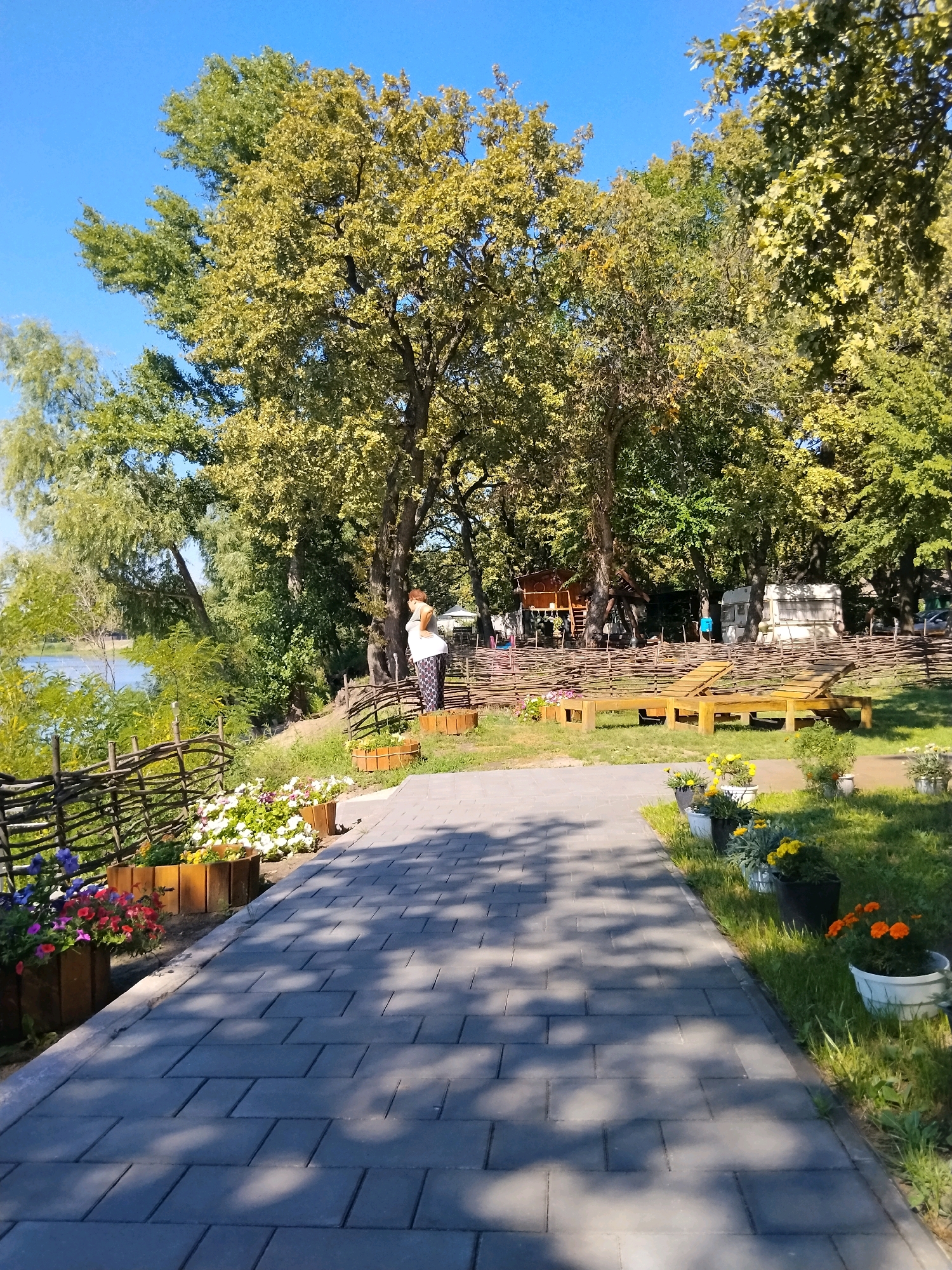 The image depicts a paved pathway leading into a park-like area in Kurylivka, Ukraine. The pathway is made of grey rectangular bricks and is flanked by planters filled with colorful flowers. To the left of the pathway, a body of water is visible, bordered by a woven fence and lush green foliage. Further into the park, a woman stands near a wooden fence, wearing a white top and patterned pants. Beyond her, several trees with vibrant green and yellow leaves dominate the landscape. In the background, a wooden playhouse structure and a camper van are visible, suggesting a recreational or camping area. The overall environment is sunny and appears to be a pleasant outdoor space.