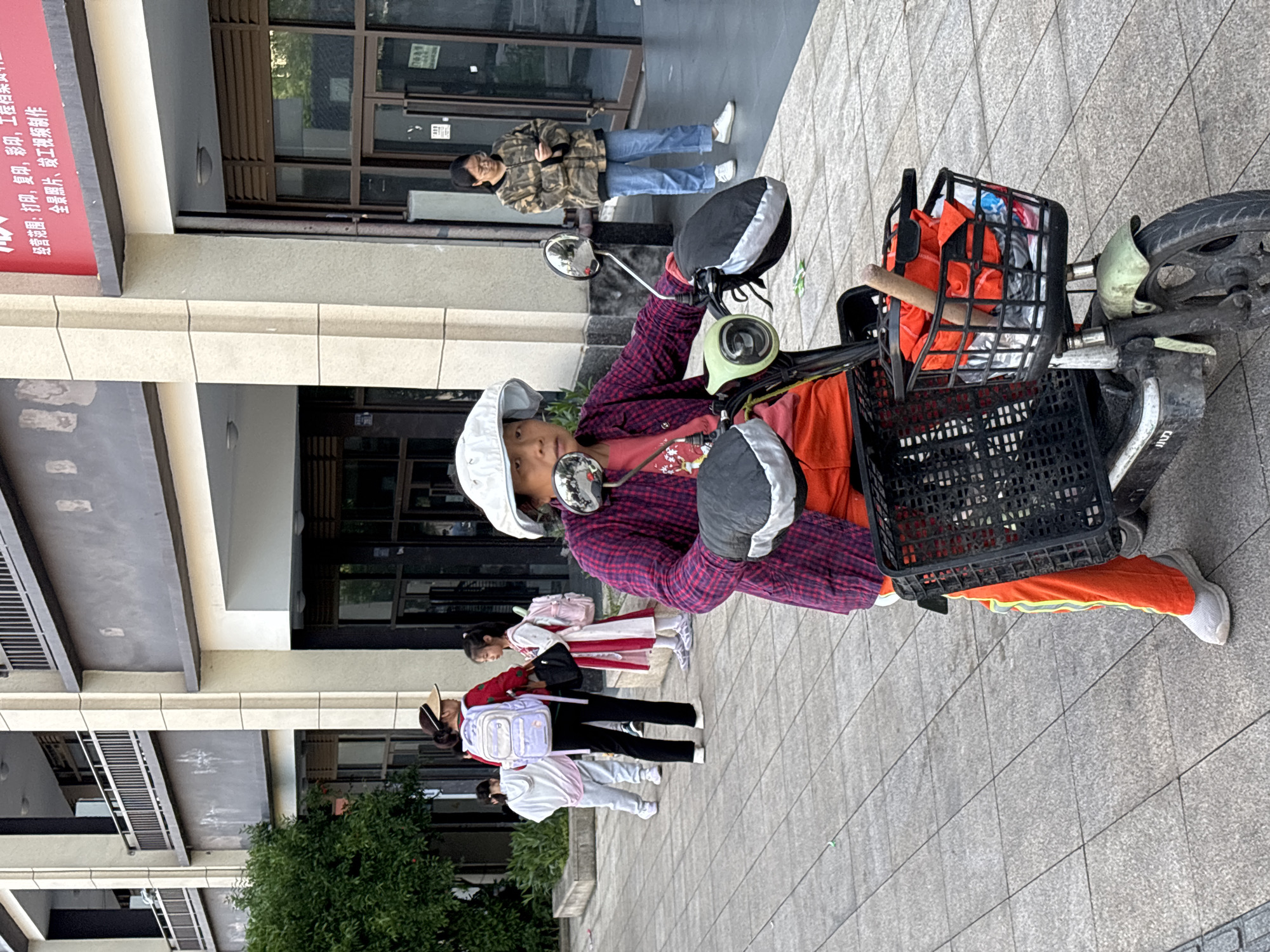 This outdoor street scene in Jiangjin, China, depicts a casual daytime moment. The primary subject is a young person, likely a child or pre-teen, with short dark hair, wearing a white cap, a red and black plaid shirt, and bright orange pants with a yellow reflective stripe. They are seated on an electric scooter, looking directly at the camera with a neutral expression. The scooter features rearview mirrors, grey and black handlebar covers, and two front baskets; the top basket is filled with orange fabric items and other clutter.
In the background, several other individuals are visible. A person in a camouflage jacket and blue jeans stands near a building entrance, also looking towards the camera. Further down the sidewalk, a group of three younger girls, possibly students, are seen. One girl wears a red and white dress (potentially a school uniform) and a light backpack, while another wears dark clothes and a light backpack.
The setting includes a modern building with large windows and a light-colored facade. A red sign on the building displays Chinese characters, detailing business services such as "printing, photocopying, color printing, engineering documents," and "panoramic photos, completion video production." The ground is a grey tiled pavement, and some green bushes are visible in the lower left corner, adding a touch of nature to the urban environment.