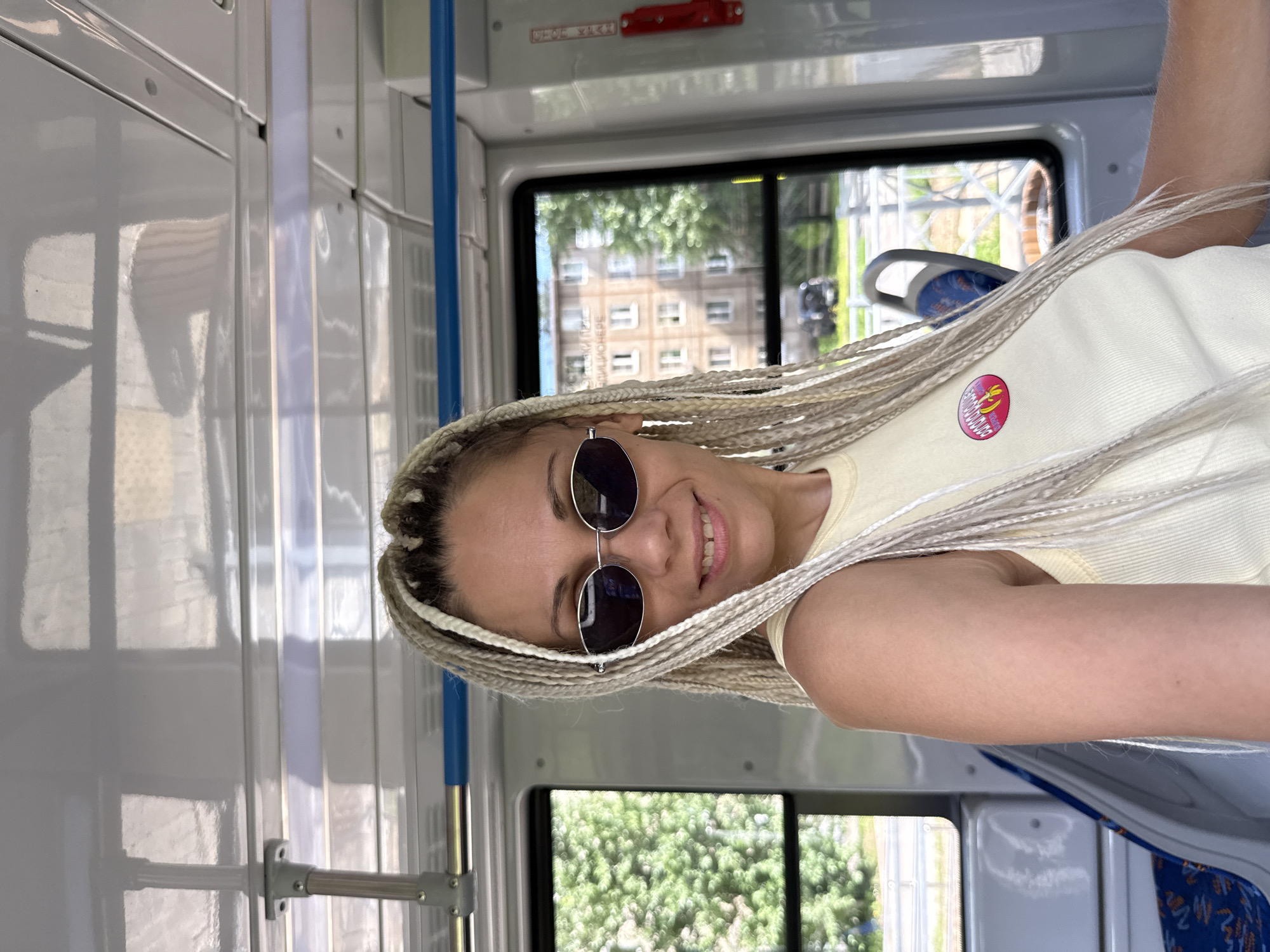 A woman with blonde braided hair and sunglasses smiles while taking a selfie on a bus in Rusanovo, Russia. She is wearing a light yellow tank top with a circular pink and yellow pin on the left side of her chest. The interior of the bus is visible, with windows showing trees and buildings outside. The seats of the bus have blue and orange patterns. The lighting suggests it is daytime.