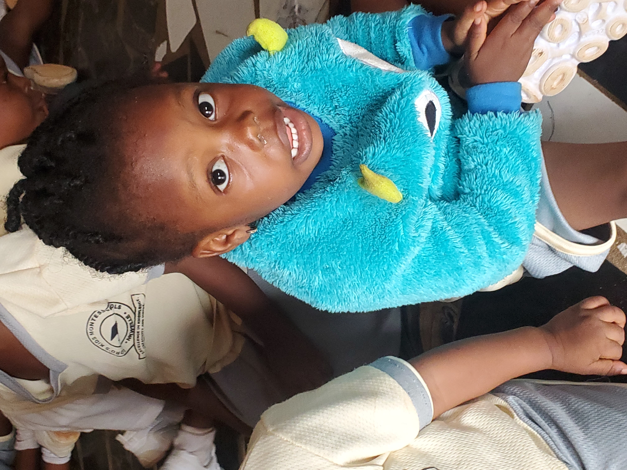 A young child with dark skin and black braided hair is looking up at the camera and smiling. The child is wearing a turquoise, fuzzy monster-themed hooded sweatshirt with yellow spikes. They are seated amidst a group of other children who are wearing similar light yellow shirts with a dark grey trim, which bear a logo for "R.O.L.S Montessori Kids" of Ukwu-Abwa, Nigeria. Some children are partly visible in the frame, suggesting an indoor group setting, possibly a school or play area. The focus is on the smiling child.
