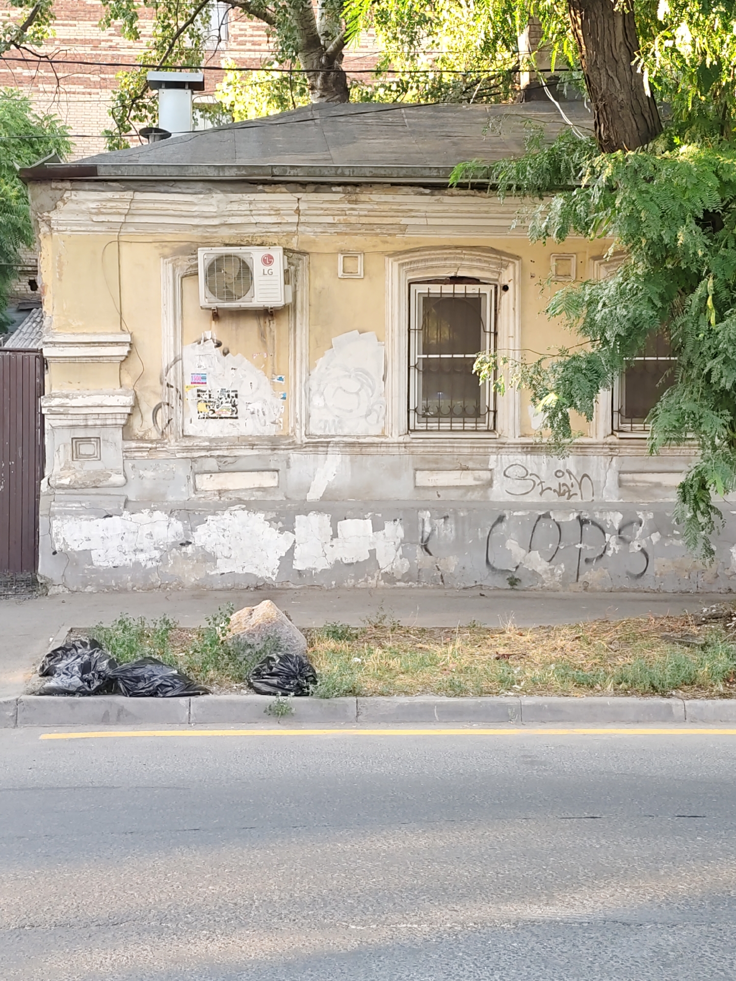 An exterior, eye-level, medium shot captures the facade of a weathered, two-story building, possibly an old house or apartment building, with evidence of decay and graffiti. The building's yellow exterior paint is peeling in many areas, revealing patches of gray plaster and brick underneath. Ornate architectural details, such as decorative moldings above the windows and pilasters on the sides, are visible but show signs of wear.
On the upper portion of the building, an outdoor air conditioning unit manufactured by LG is mounted to the wall. The windows on the facade are covered with metal bars, indicating security measures. Peeling posters and what appear to be other remnants of graffiti are scattered across the walls, particularly around the windows. Lower on the building, more prominent graffiti is visible, including the word "COPS" spray-painted in large, black letters.
In the foreground, a strip of dry, patchy grass and weeds runs along the base of the building, separated from a paved street by a concrete curb. Three black trash bags are piled haphazardly on the grassy area near the curb, along with a large, rough-looking boulder. A bright yellow line is painted on the asphalt street, suggesting a road or parking area. The scene is illuminated by natural sunlight, casting shadows and highlighting the textures of the worn building and surrounding environment. The overall impression is one of urban neglect and disrepair. The location is identified as Rostov-on-Don, Russia, based on the provided context.