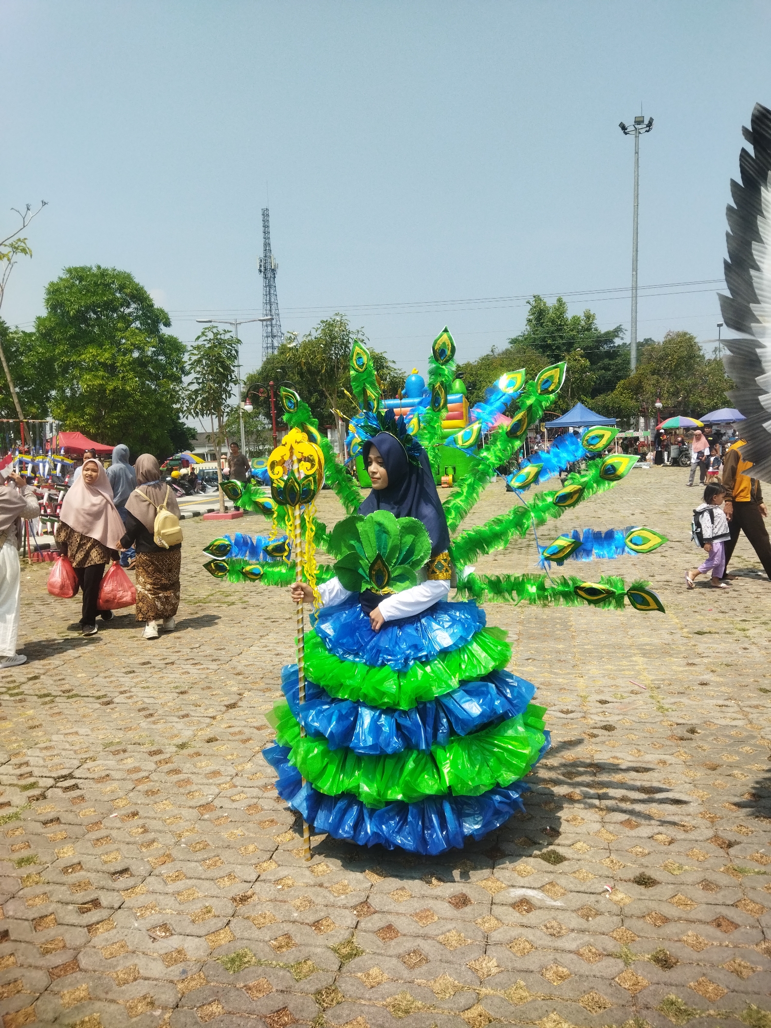 In the city of Wonogiri, Indonesia, a young woman is dressed in an elaborate peacock costume made of green and blue plastic bags, decorated with peacock feather motifs. She stands in an outdoor plaza paved with interlocking bricks, holding a yellow staff adorned with peacock feather designs. The background shows a crowd of people browsing stalls under tents, with trees and a telecommunication tower visible against a clear blue sky. A tall lamppost stands to the right of the frame.