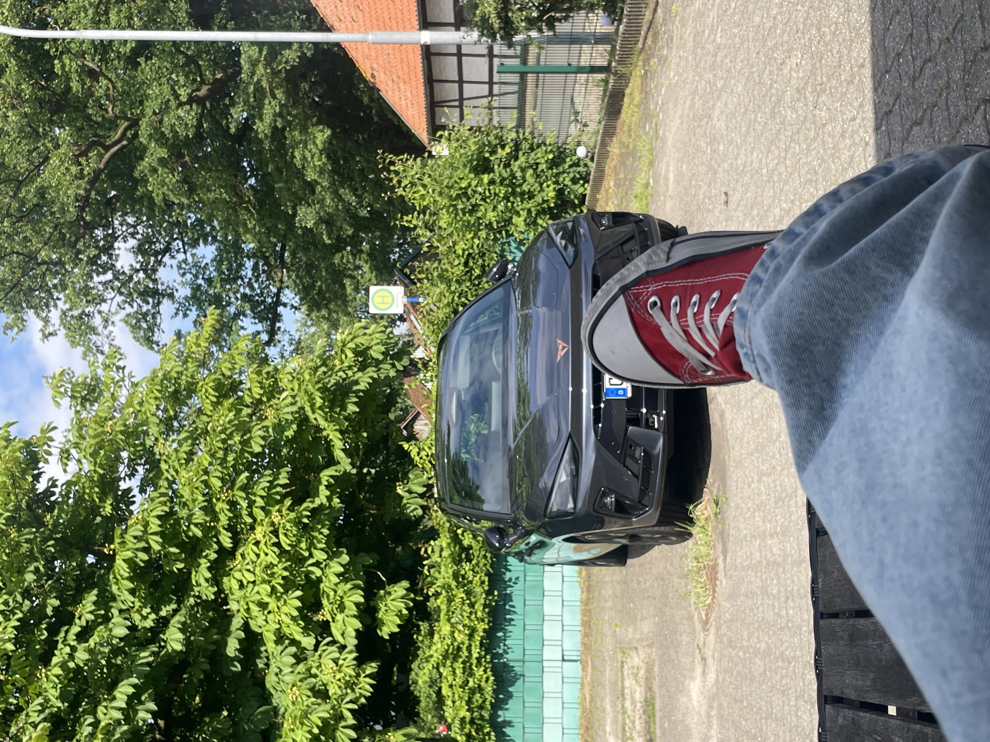 A casual outdoor shot features a person's leg, clad in light blue jeans and a red canvas shoe, casually extended towards a dark gray car parked on a paved area. The car is positioned adjacent to a green fence and lush greenery. A street sign with a "H" symbol is visible amongst the foliage, and the background includes a house with a red tile roof and traditional half-timbered detailing. The image appears to be taken at ground level and slightly angled. The setting is Wienhausen, Germany.