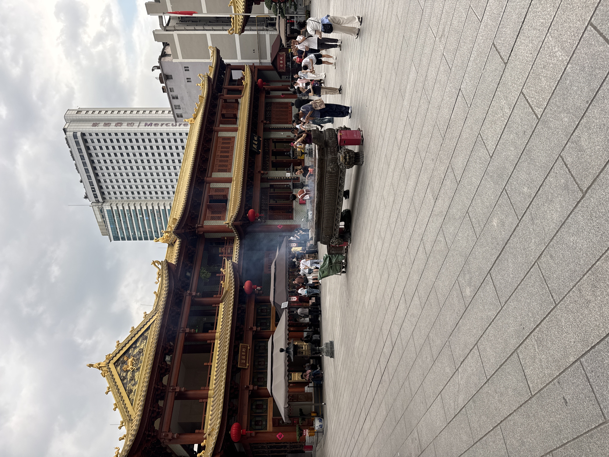 The image shows a temple in Yuexiu District, China. The temple is built with red and gold architecture, and there are people in front of it. There are also some modern buildings and a cloudy sky in the background. People are burning incense in front of the temple.