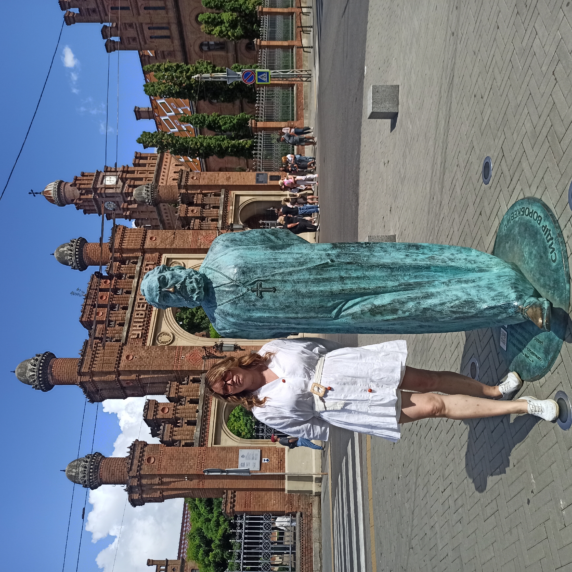 A woman in a white dress stands to the left of a large bronze statue of a bearded man wearing a robe and cross pendant, in front of the main entrance archway of Chernivtsi National University (formerly Residence of Bukovinian and Dalmatian Metropolitans) in Chernivtsi, Ukraine. The university building is constructed of red brick with distinctive turrets and a clock tower. The statue is positioned on a circular base with Cyrillic writing. A crosswalk is visible in the foreground. Several people are walking in the background behind a decorative fence. The sky is blue with scattered white clouds.