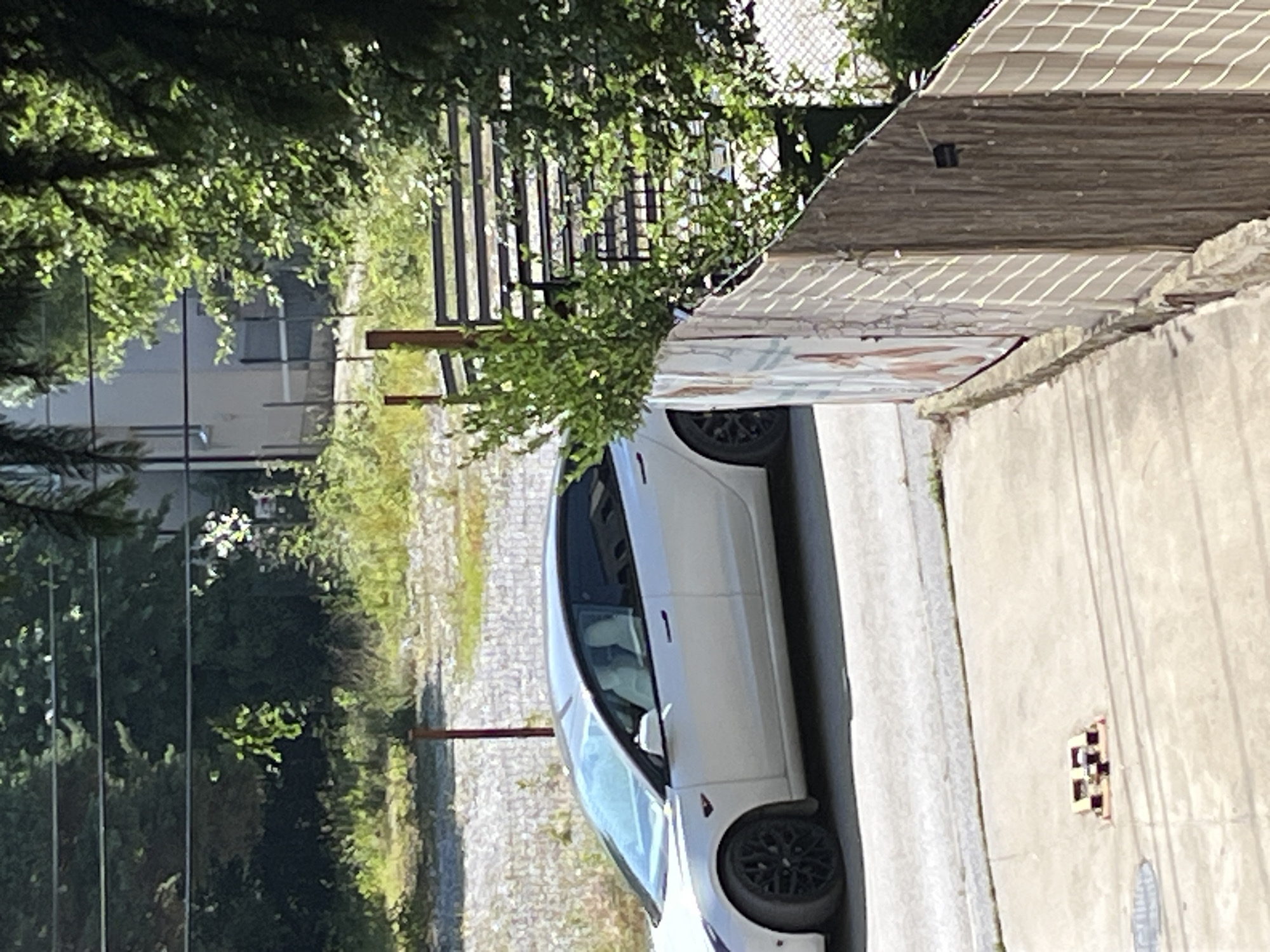 A white Tesla Model 3 is parked on a paved surface next to a chain-link fence with wooden planks and a white tarp. The car is facing left, with its front wheel turned inward. Behind the car, there is an overgrown area with weeds and trees, and a building with a light-colored facade is visible in the distance. A black metal fence separates the foreground from the background. The scene appears to be in an urban or suburban setting.