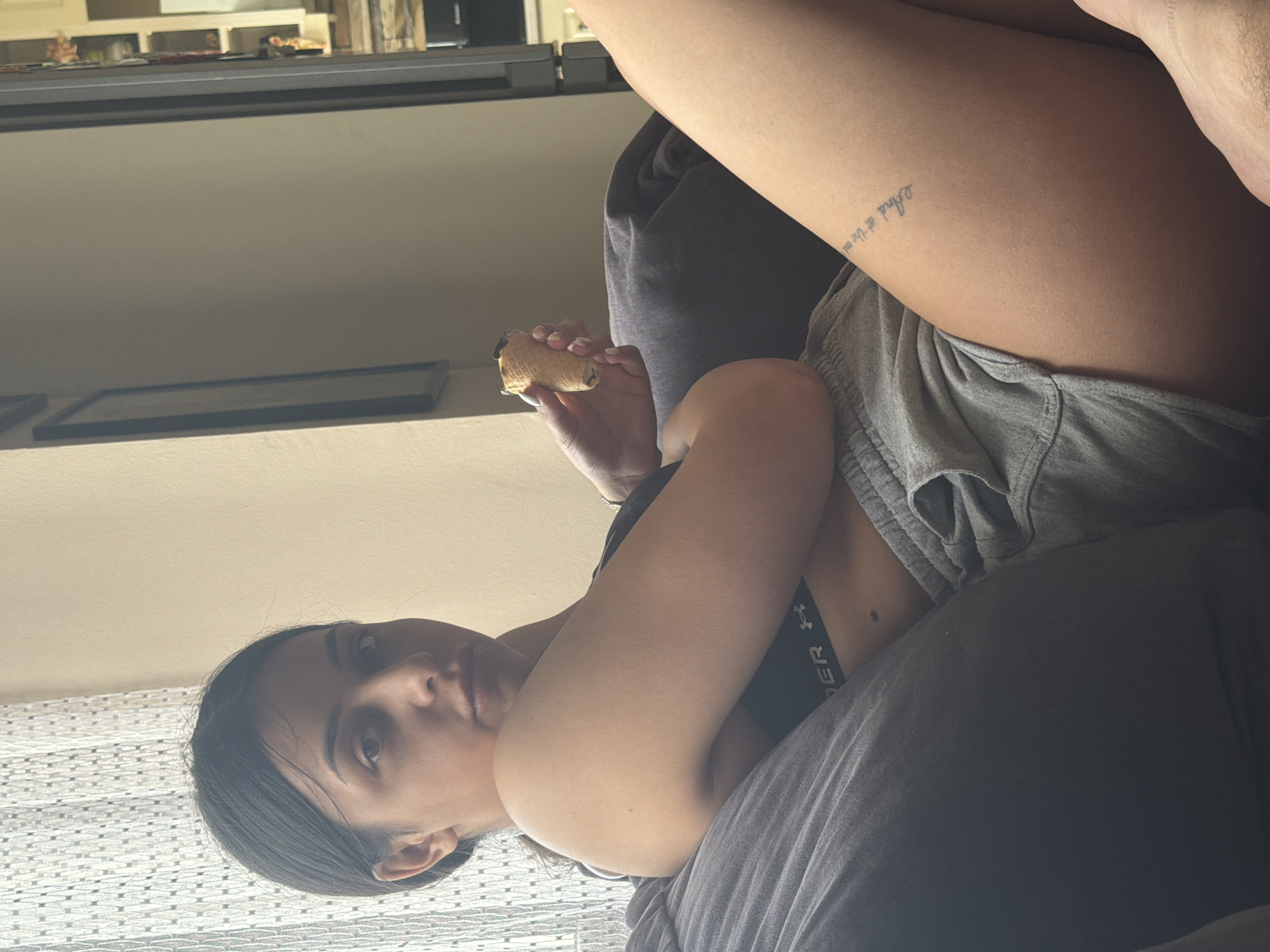 The image, rotated 90 degrees clockwise, captures a young woman reclining casually indoors, likely on a dark grey sofa or bed. She has dark hair and is looking directly towards the camera with a neutral expression. She is wearing a black bra or sports bra, with the brand name "Under Armour" visible on the band, and light grey shorts.
In her right hand, she holds a light-colored, rolled pastry, possibly a cannoli or similar sweet, with a dark filling, suggesting she is eating it. A small, faint text tattoo is visible on her upper left thigh, reading "And at the en".
The background features a light-colored wall with a window covered by white horizontal blinds or curtains on the left side of the frame (top in the original orientation). Below the window, a rectangular framed object rests on a surface, and further back, another shelf or ledge holds a few indistinct items. The overall lighting suggests it is daytime. The setting appears to be a domestic space. The provided location context is Shumen, Bulgaria.
