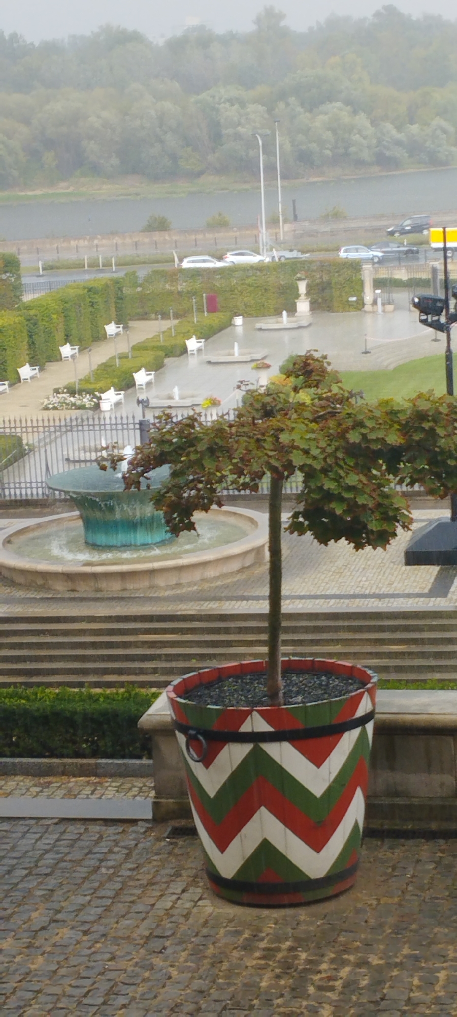 The image captures a slightly misty outdoor scene in Warsaw, Poland. In the foreground, a tree is planted in a large, cylindrical pot adorned with a chevron pattern of white, red, and green. The pot has black bands and ring handles on the sides. Behind the pot, there are stone steps leading up to a terrace. Further back, a fountain with a teal-colored basin is visible, with water circulating. Beyond the fountain, there is a neatly manicured garden with rows of white benches and hedges, interspersed with smaller fountains. A metal fence encloses this garden area. In the background, a body of water, likely the Vistula River, is seen, with a wooded embankment on the opposite shore. Several cars are parked or driving on a road that runs parallel to the river, with streetlights and lampposts visible. The overall atmosphere suggests a public park or a well-maintained garden area within the city.