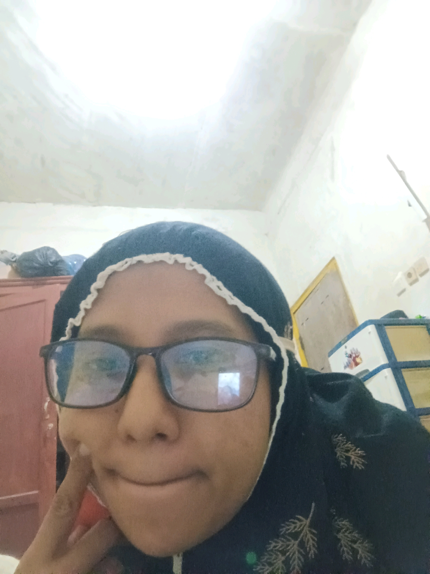 A young woman, wearing glasses and a dark headscarf with white trim, is taking a selfie indoors. She is leaning her cheek on her hand and appears to be in a bedroom or living space. In the background, a red cabinet or dresser is visible, along with a white plastic storage unit with blue trim. The walls are textured and painted white, and there are electrical outlets on the wall. The overall scene suggests a casual, personal moment captured within a home environment in the city of Sarangan, Indonesia.