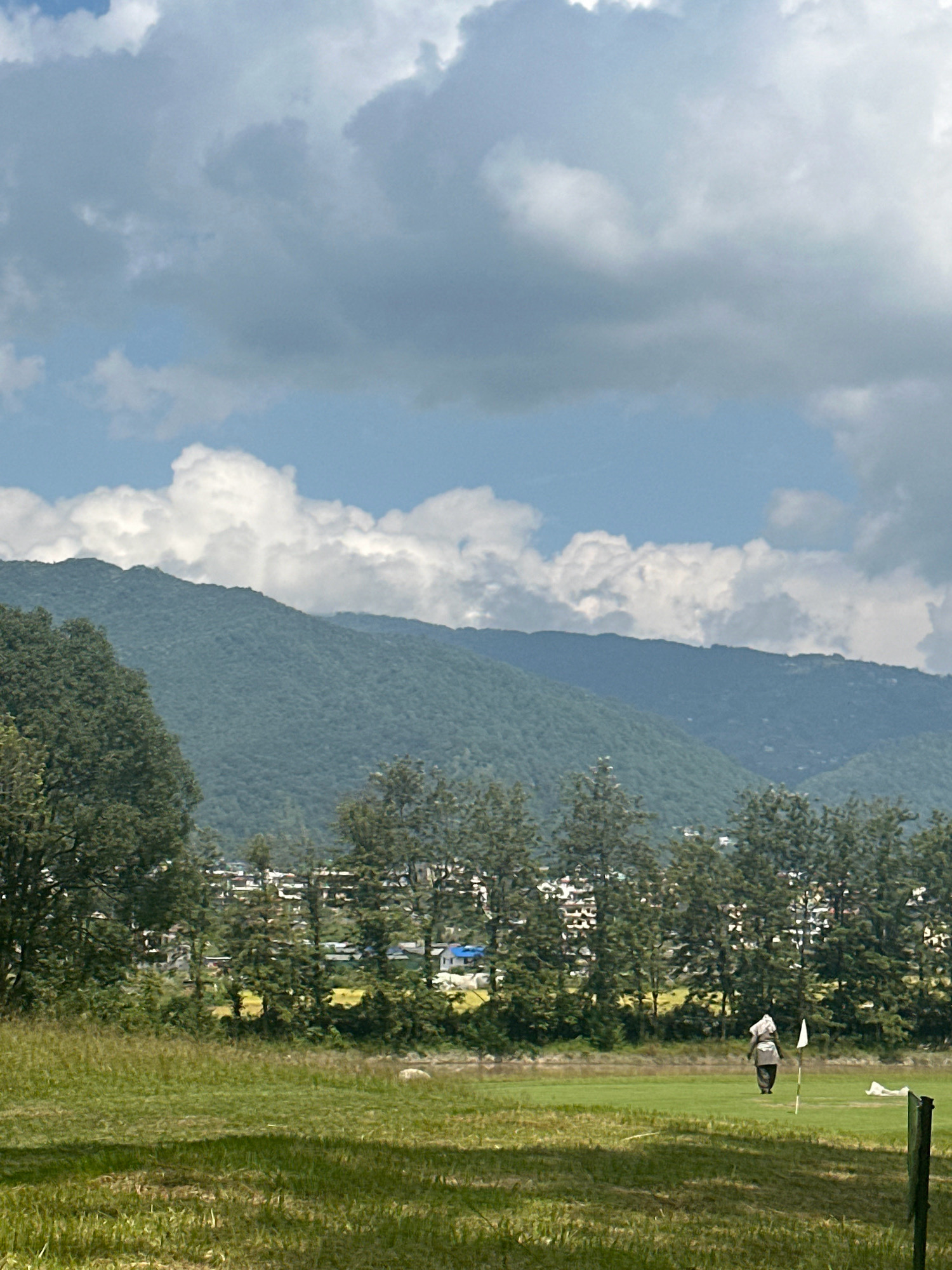 A medium shot captures a golf course scene in Gokarneshwar, Nepal, under a sky filled with dramatic clouds. In the foreground, a lush green lawn is partially covered by shadows, suggesting the presence of trees or structures not fully in frame. Further back, a golfer, dressed in a white head covering and dark clothing, walks away from the viewer towards a golf flag planted in the ground. To the right of the flag, a white object lies on the grass, possibly a discarded piece of clothing or golf equipment. Beyond the golf course, a line of trees forms a natural barrier, behind which a cluster of buildings with white roofs and some blue accents are visible, indicating a populated area. The background is dominated by rolling, densely forested hills that stretch towards the horizon, their slopes partially obscured by the dramatic clouds.