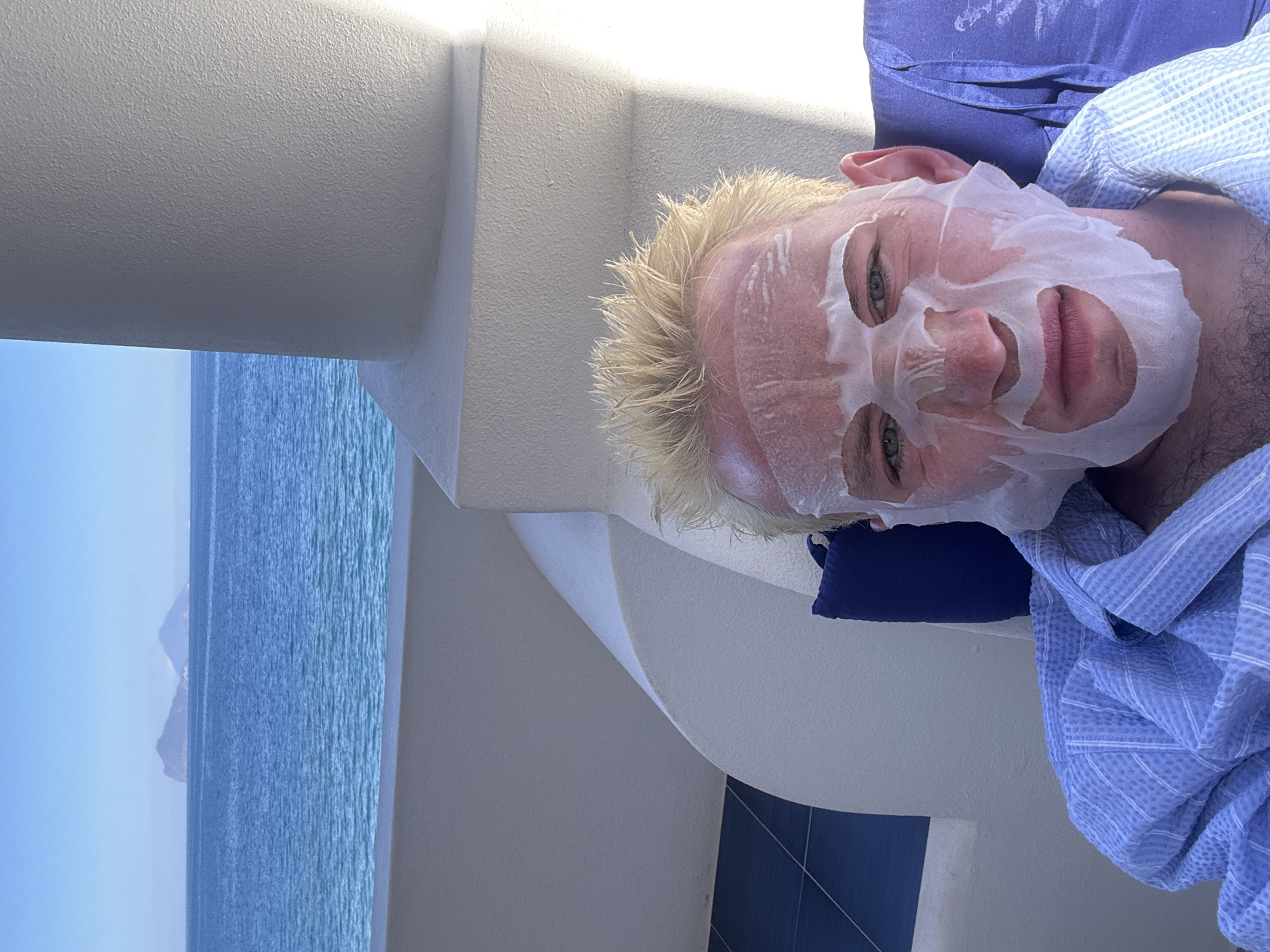 A person is seen from a slightly elevated angle, lying outdoors on what appears to be a balcony or patio. The individual has blond, spiky hair and is wearing a white sheet mask that covers most of their face, leaving the eyes, nose, and mouth exposed. Their eyes are open and looking forward. They are partially covered by a light blue and white striped blanket or robe, and a dark blue pillow is visible behind their head.
The setting features modern, light-colored architectural elements, including a white, textured wall, a smooth white ceiling, and a curved white railing that forms part of the balcony structure. In the background, a vast, calm body of clear blue water (the sea) stretches out under a bright, clear sky. A faint, distant landmass or mountain is visible on the horizon, contributing to the serene coastal view. The overall impression is one of relaxation in a sunny, outdoor environment, likely a resort or hotel balcony in Lipari, Italy, given the context. The time of day appears to be bright daytime, with excellent visibility. No text is visible in the image.