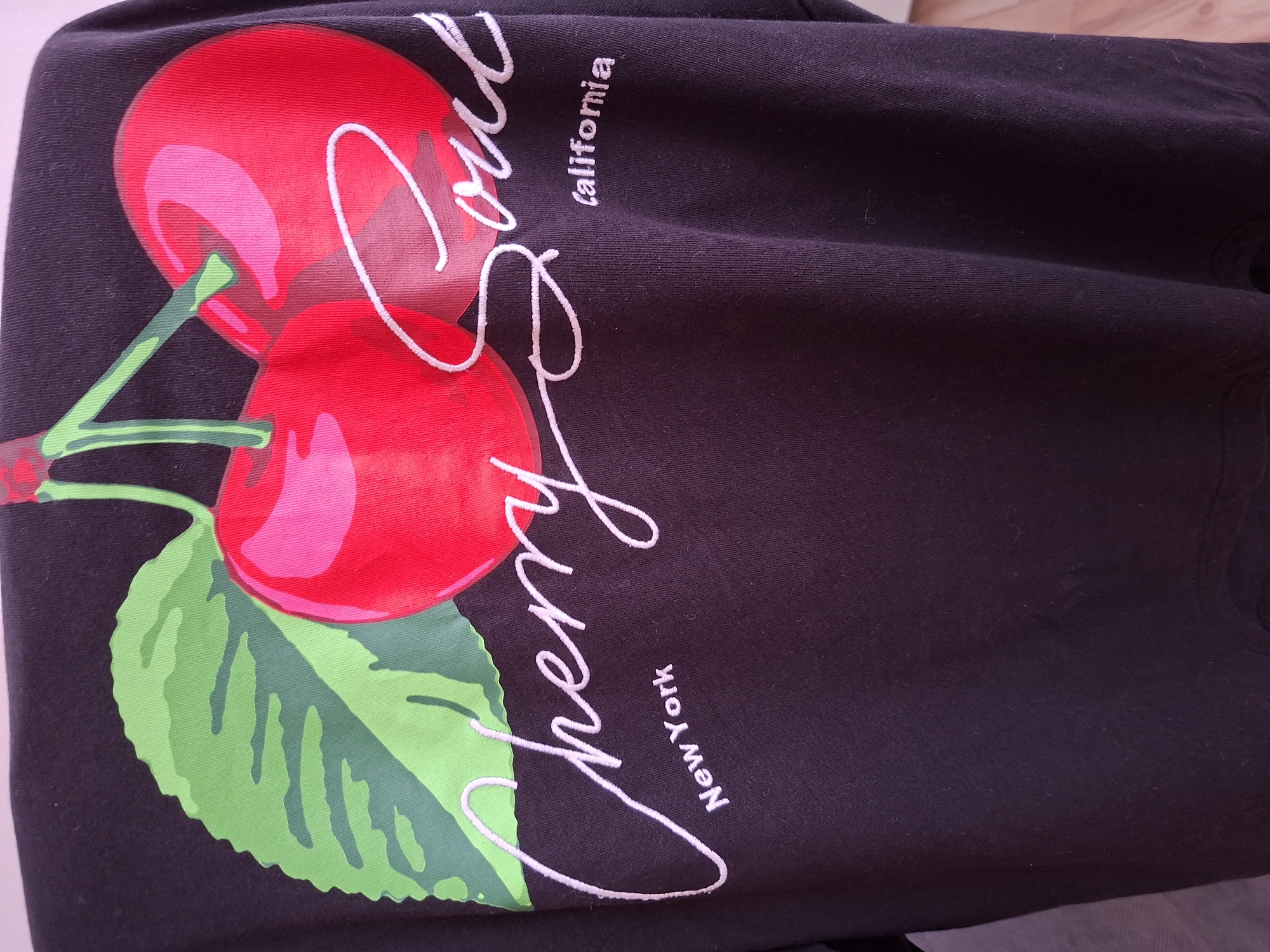 The media file displays a dark purple or black piece of clothing, likely a t-shirt or sweatshirt, laid flat. The main subject is a large, vibrant graphic print featuring two glossy red cherries with bright green stems and a prominent green leaf. The cherries have highlights, giving them a three-dimensional, almost juicy appearance.
Overlaid on the design is white text. The central text is "Cherry Soul" written in a flowing cursive script. Above the word "Soul," the smaller text "California" is printed vertically. Below the word "Cherry," the smaller text "New York" is also printed vertically, but oriented in the opposite direction, suggesting the garment is rotated.
The fabric of the clothing appears soft and has subtle folds and creases, indicating it's not stretched taut. There are a few small fibers or lint visible on the dark material in some areas. The overall setting is indoors, with the garment possibly laid on a light-colored surface, part of which is visible in some cropped hints. There is no specific activity taking place, as it's a static image of an item of clothing. The location context is Plovdiv, Bulgaria.