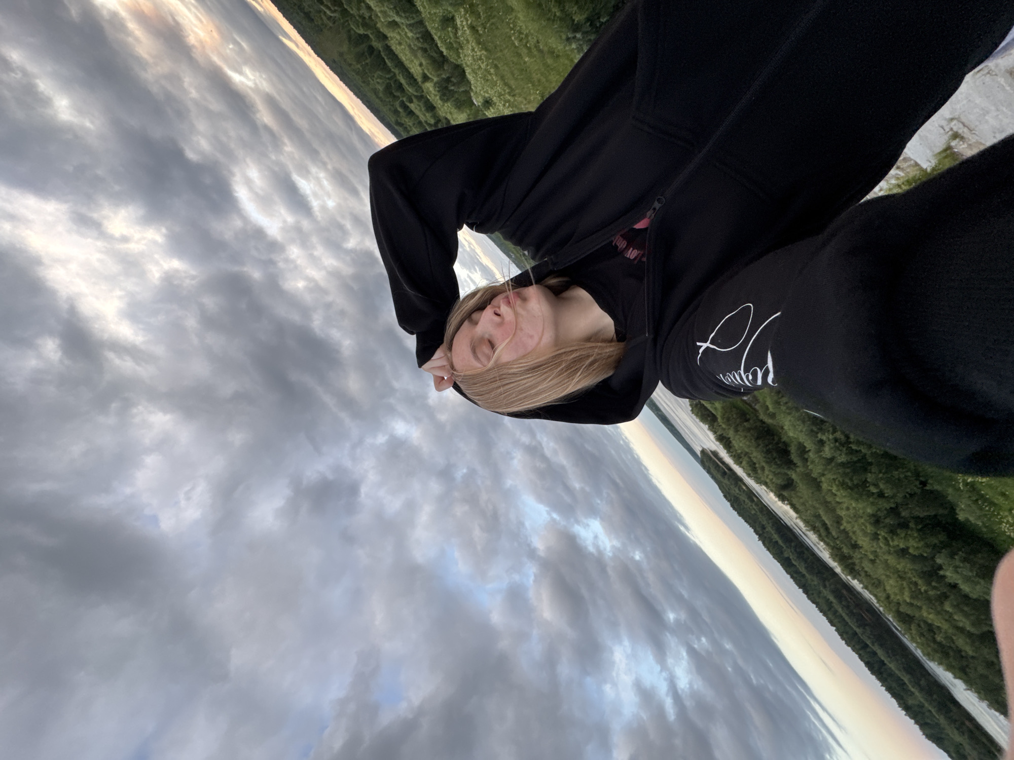 A young, blonde-haired person in a black hoodie and t-shirt is taking a selfie outdoors. Their right hand is raised, resting on their head, and their lips are puckered as if blowing a kiss. The background shows a cloudy sky and a lush green landscape with trees and a body of water, possibly a river. The image is taken from a high vantage point, suggesting the person is on a hill or elevated area. The location is identified as Ryabinino, Russia.
