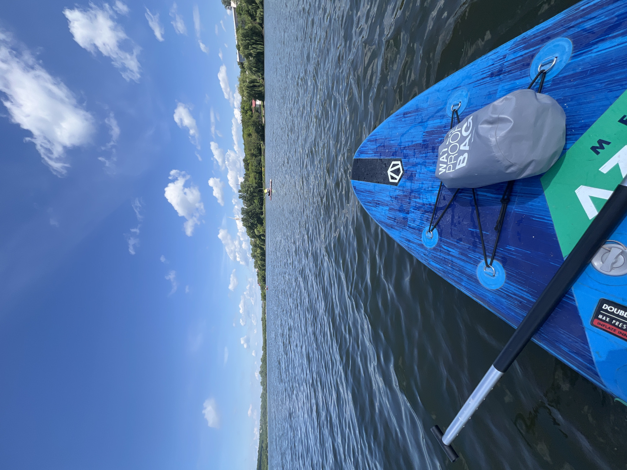An individual is paddleboarding on a body of water with a blue paddleboard in the foreground. The paddleboard has a gray waterproof bag secured to it. The water is dark blue with moderate ripples. In the background, there is a shoreline with lush green trees and some buildings. The sky is bright blue with scattered white clouds. The location is Ternopil, Ukraine, likely on Ternopil Lake.