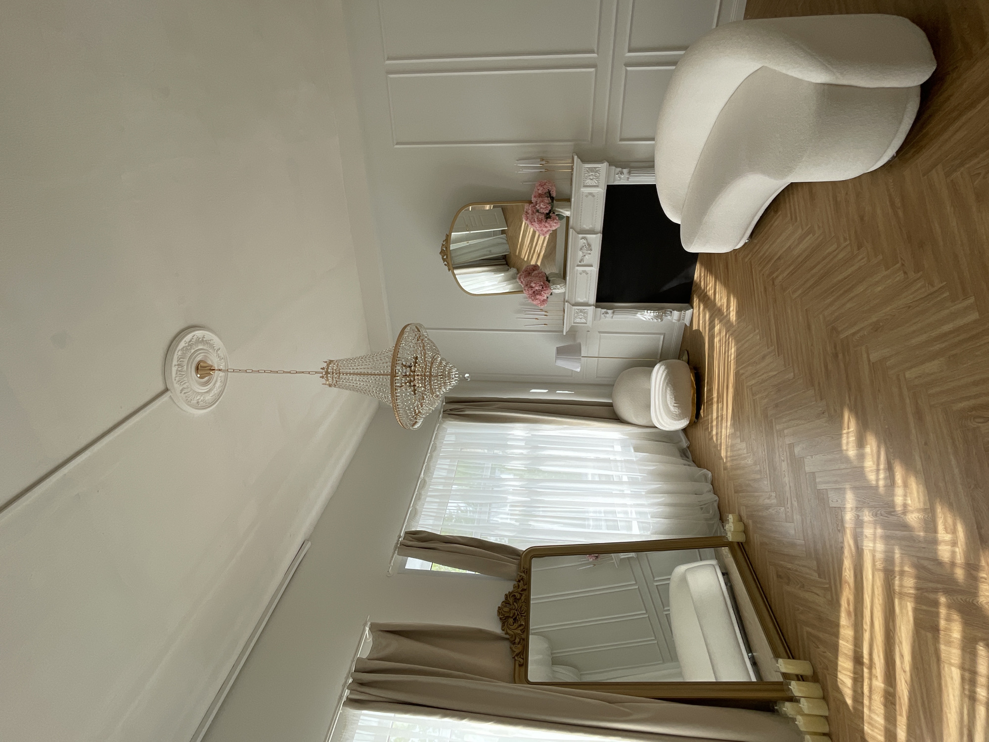 The image shows the interior of a room in what appears to be a domestic setting, possibly a residence, with elements that evoke a luxurious or decorative style. A chandelier hangs from the ceiling, and a fireplace mantle with a black inset is visible against the wall. A white, organically shaped chair stands near the fireplace. A floor-length mirror, in a gold frame, reflects a portion of the room, including a white cushion. Light streams into the room through window draped in beige curtains. The flooring has a parquet pattern. The location, Kazan, Russia, cannot be confirmed from the image alone.