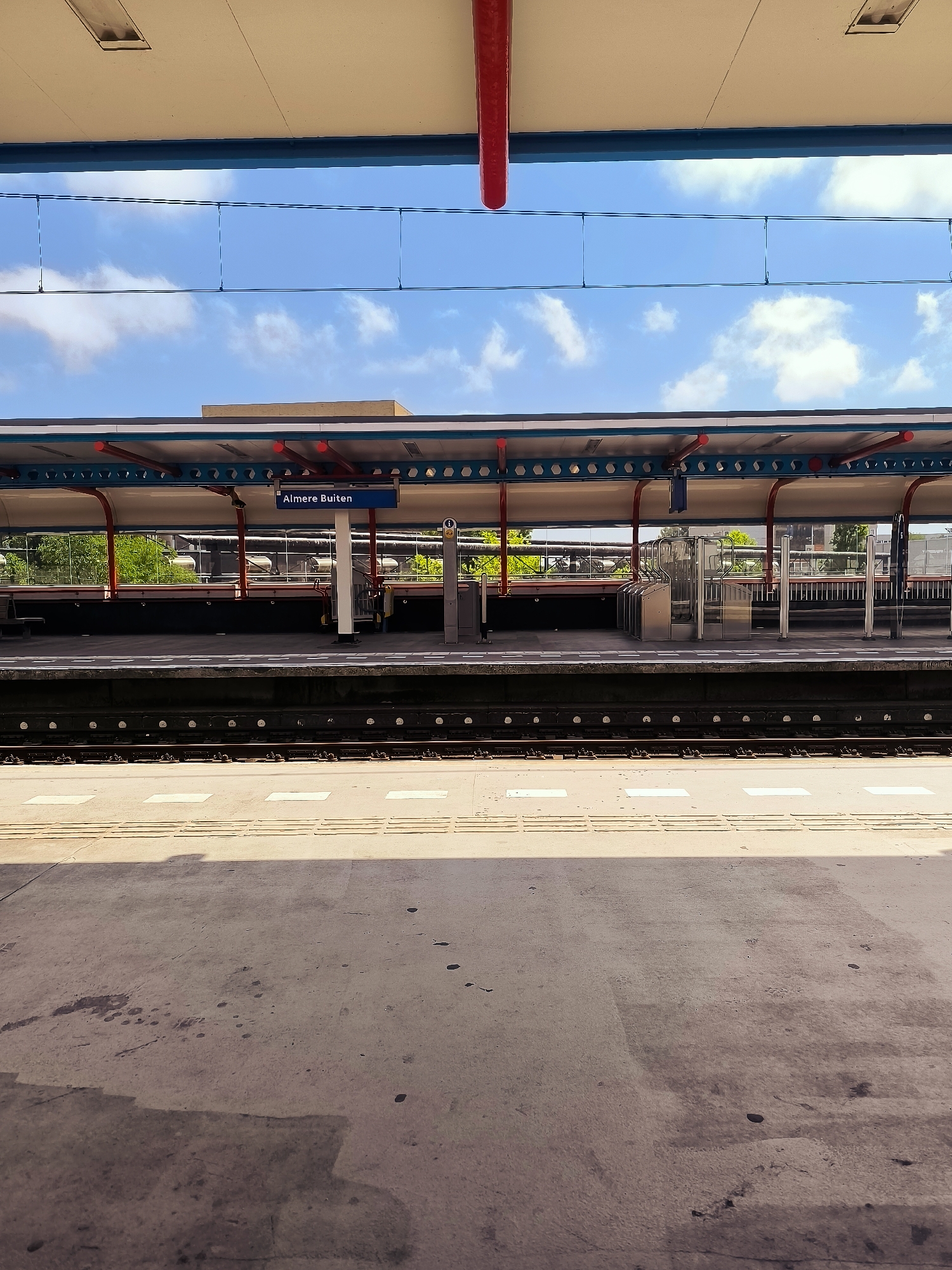 This image depicts the platform of Almere Buiten train station in Almere, Netherlands. The platform is made of concrete and features tactile paving for visually impaired individuals. A covered waiting area with a blue and red trim runs along the platform, supported by red pillars. A blue sign indicates "Almere Buiten". Behind the platform, trees and buildings are visible. The sky above is blue with scattered clouds. The scene is empty of people.