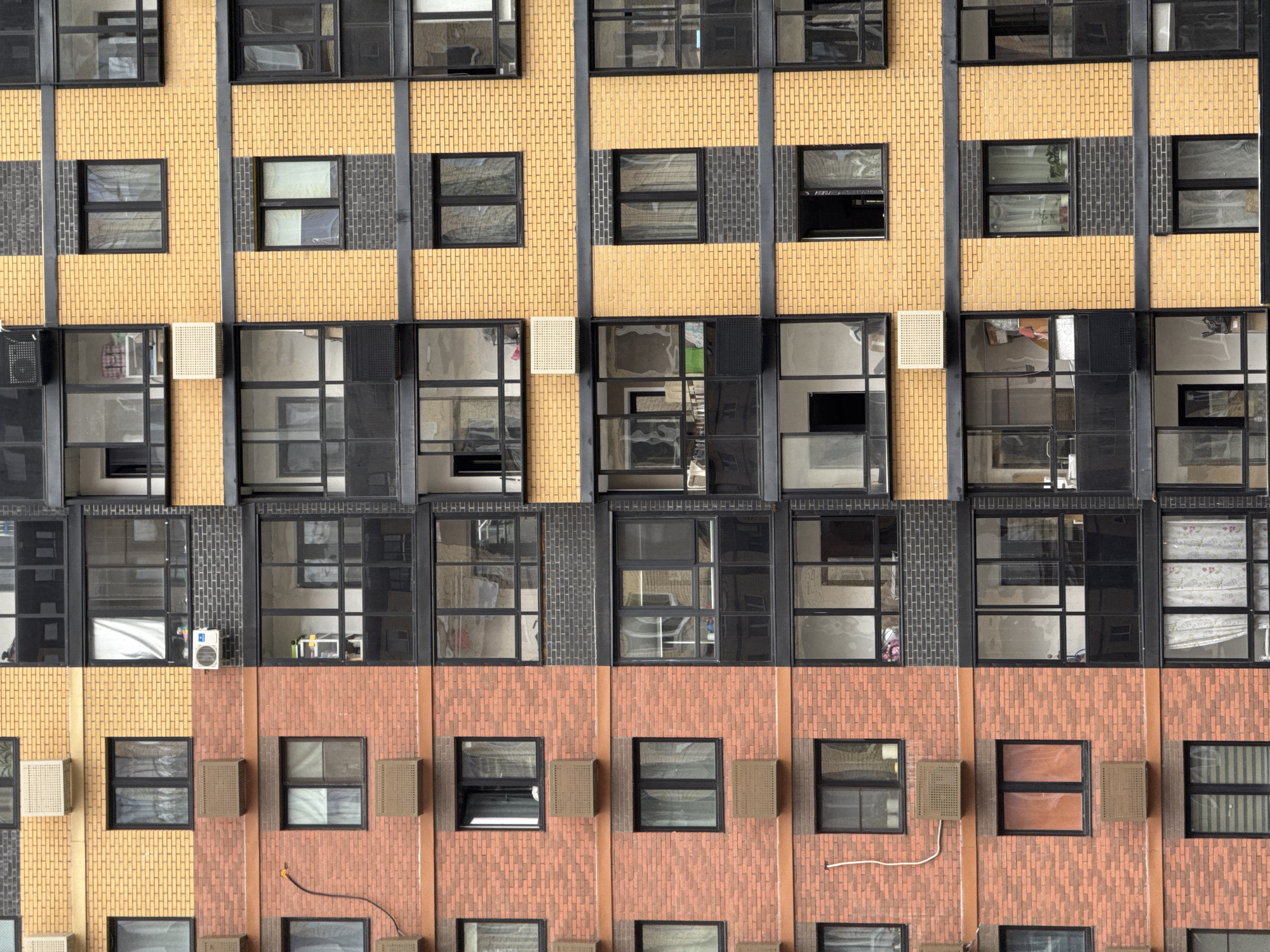 A section of a multi-story apartment building in Mirny, Russia, features a split facade design. The left side is clad in red brick, while the right side is a combination of yellow brick and dark grey brick. The building has numerous windows and balconies, many with black frames. Several air conditioning units are visible mounted on the exterior walls. The windows reveal glimpses of the interiors, showing curtains and some household items. The overall impression is that of a modern residential structure in an urban environment.