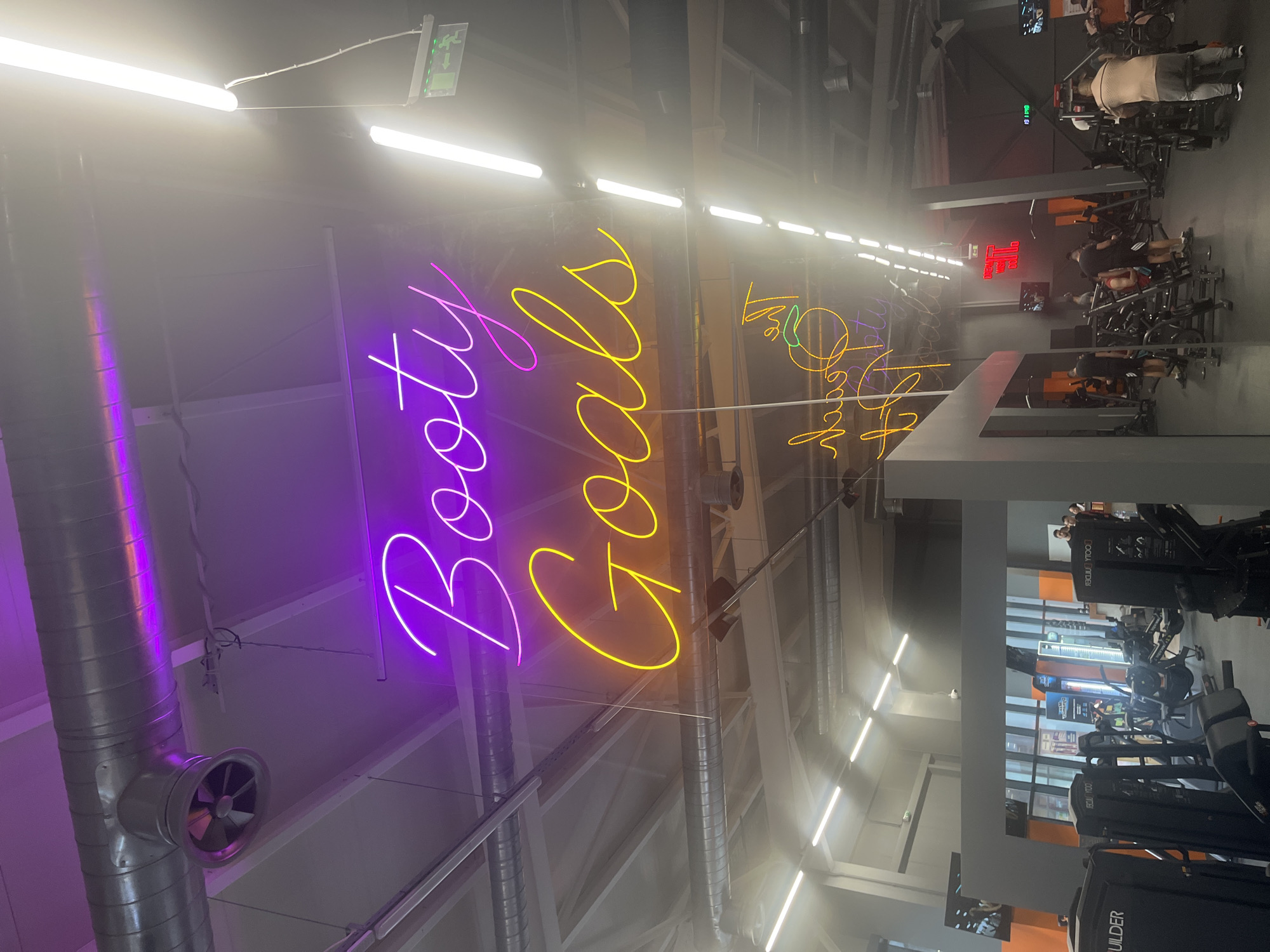 The image depicts the interior of a gym, potentially in Plovdiv, Bulgaria. Neon signs reading "Booty Goals" in purple and yellow, and "Go Big or Go Home" in orange are visible on the ceiling. Exercise equipment, including rowing machines, are arranged in rows. People are present, some using the machines. Fluorescent lighting illuminates the space. Ductwork runs along the ceiling. A digital clock displays the time.