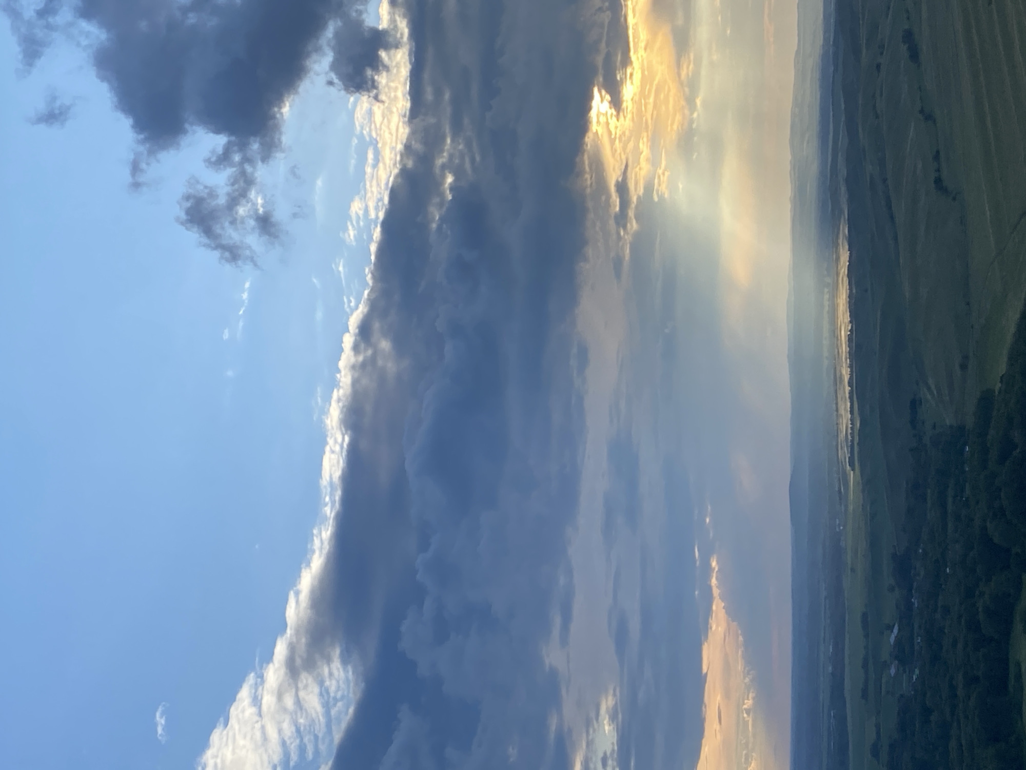 This is an aerial, wide-angle photograph of a rural landscape at sunset. The sky is a dominant feature, filled with dramatic, layered clouds. The upper portion of the sky is a clear, vibrant blue, transitioning to darker, textured gray clouds that stretch across the frame. Shafts of golden sunlight break through the clouds, illuminating patches of the landscape below. The middle ground features rolling hills covered in a patchwork of green fields and scattered trees. In the distance, a hazy horizon is visible, with faint outlines of more hills. The foreground shows a dense area of dark green trees. There are no people or specific structures clearly identifiable in the image, suggesting a natural, undeveloped setting. The scene evokes a sense of natural beauty and the grandeur of the sky at dusk.