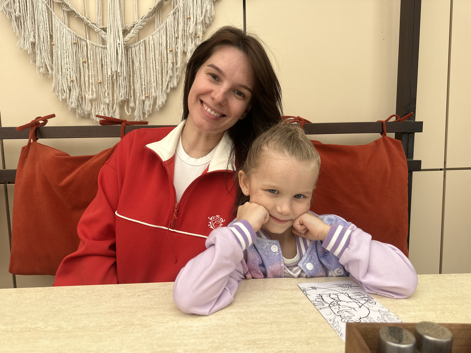 In the city of Chaikovsky, Russia, an image captures a woman and a young girl seated at a table within an indoor establishment. The woman, with dark hair and a bright smile, wears a red sweater with a white collar over a white t-shirt. The young girl, with her blonde hair pulled up, rests her chin on her hands, her elbows on the table. She is wearing a light purple jacket over a white shirt. A coloring sheet lies on the table in front of her. Behind them is a cushioned bench, colored a muted red, set against a wall. Above the bench is a decorative macrame wall hanging. The table is light-colored. Salt and pepper shakers can be seen in the foreground.