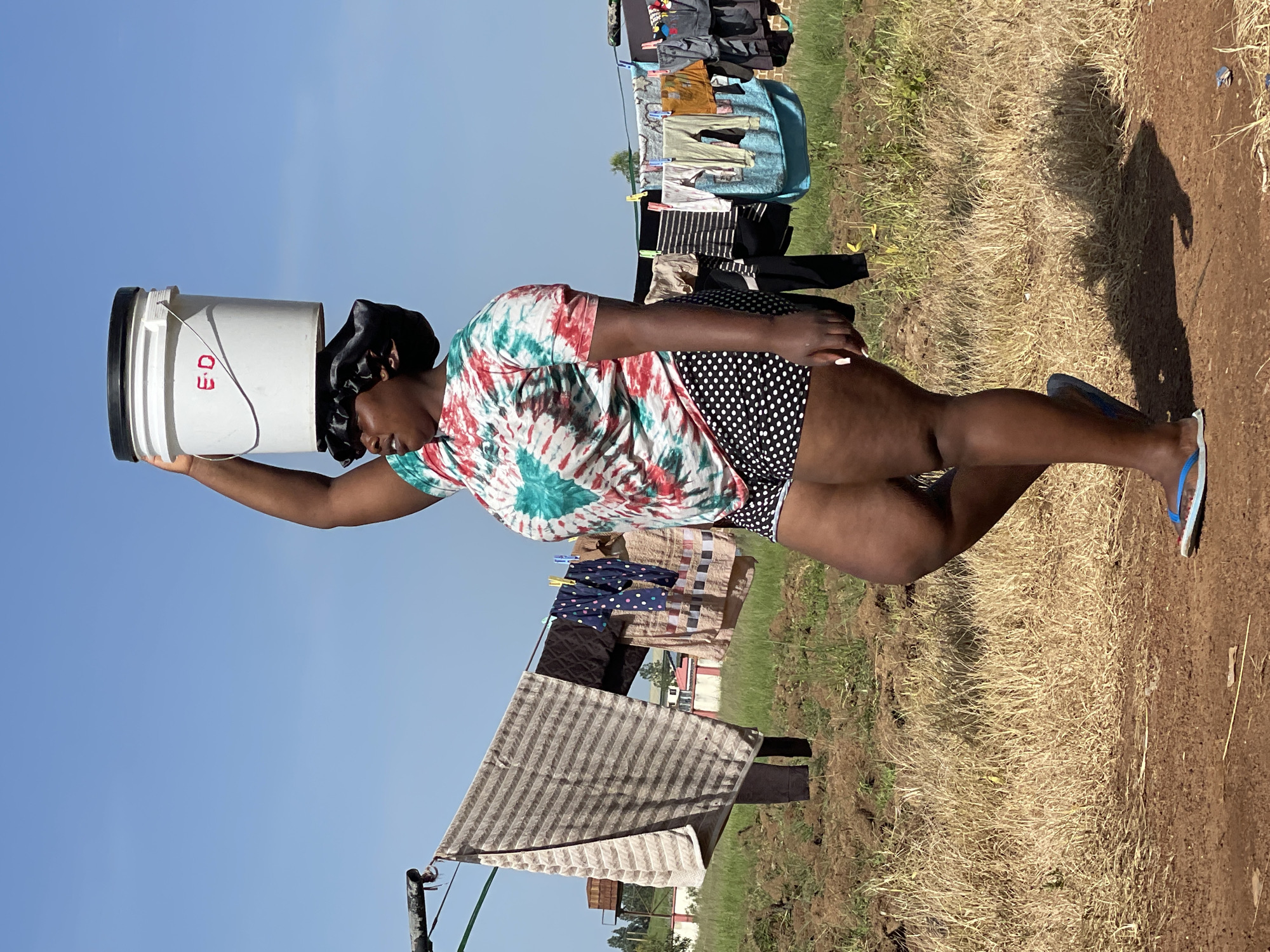 In Durumi, Nigeria, a young woman is captured in a medium shot, balancing a white bucket with a black lid on her head. She is wearing a colorful tie-dye t-shirt and black and white polka dot shorts, and a black satin bonnet. The woman is standing in an outdoor setting with a clothesline in the background, strung with various garments. The ground is a mix of dry grass and soil. The sky is clear and blue, indicating a sunny day. The background shows a few scattered buildings and trees. The woman's posture suggests she is walking or about to walk.