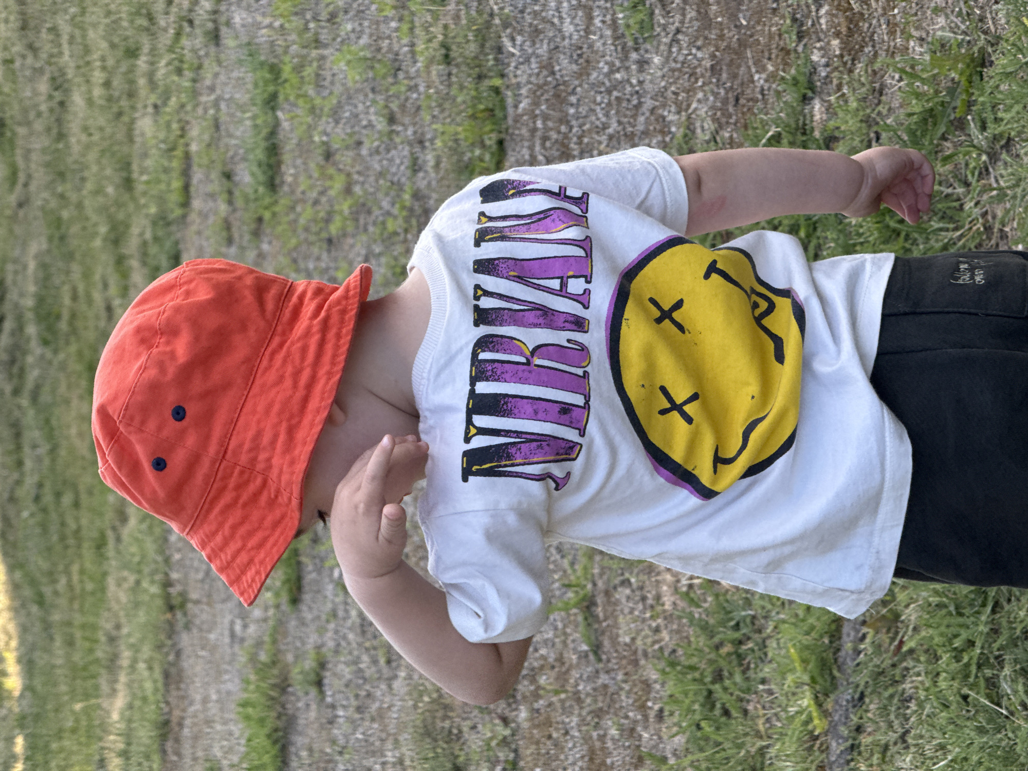 A child is wearing a white t-shirt with the word "NIRVANA" printed in purple and yellow, and a yellow smiley face with two black X's for eyes and a curved line for a mouth. The child is also wearing a bright orange bucket hat with two dark blue buttons on it. The child is standing in a grassy area with some dry weeds. The lighting suggests it is daytime, likely outdoors. The child's right arm is bent at the elbow and their hand is near their face. The scene is an outdoor setting, with a slightly blurred background of grass and possibly some dry vegetation. The image is oriented vertically.