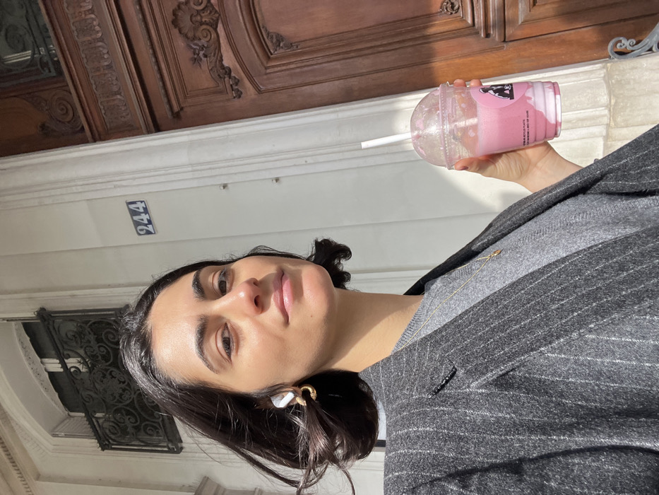 An adult female is depicted in a close-up, outdoor shot, likely a selfie, on a sunny day. She has dark, shoulder-length hair and is looking directly at the camera with a slight smile. She is wearing a grey pinstriped blazer over a grey sweater, a gold-colored necklace with a small pendant, a white wireless earbud in her left ear, and a gold-toned hoop earring in her left ear. In her right hand, she holds a clear plastic cup with a dome lid and white straw, containing a two-toned pink and white beverage. The cup displays a black logo featuring a gorilla figure and partially visible text indicating "100% RECYCLED PLASTIC."
The background reveals architectural details of a classic Parisian building. A blue and white enamel plaque with the number "244" is affixed to a white stone facade. Above this, a window with a dark, ornate wrought-iron grille is visible. To the right, a section of a large, dark brown wooden door with intricate carvings and moldings is present. The distinct architectural style and street numbering confirm the location as a street in Paris, France. Sunlight strongly illuminates the scene, casting highlights on the woman's face and hair.