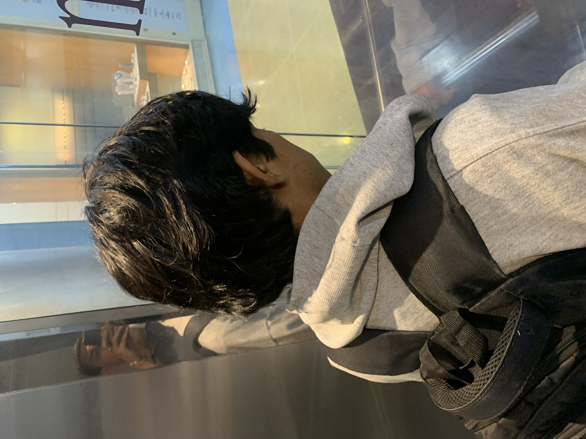 A person with short, dark hair is seen from the side and rear, wearing a gray hooded sweatshirt and a black backpack. The person is positioned in front of a reflective surface, possibly glass or polished metal, which shows a distorted reflection of their face. Through the glass, there is a glimpse of a building interior with what appears to be a display case and lettering on the top, suggesting a storefront or commercial space. Based on the prompt, this scene is located in Porlamar, Venezuela.