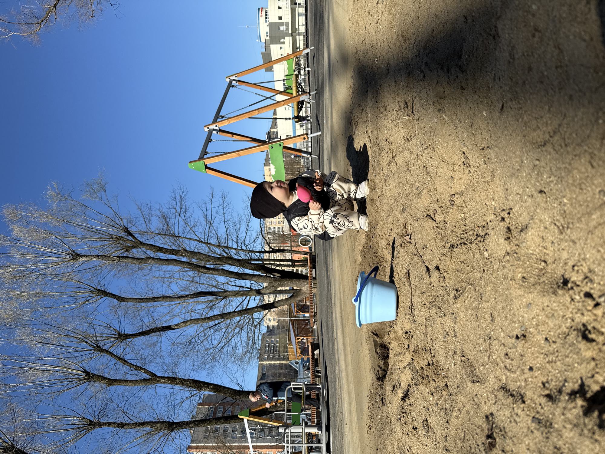 The image shows a young child sitting in a sandbox in what appears to be a playground or park. The child is wearing a black beanie, a patterned outfit (possibly a jumpsuit), and shoes, and is holding a pink object. A blue bucket sits near the child in the sand. In the background, playground equipment, including a swing set and climbing structure, is visible. Trees, mostly bare of leaves, are present, and beyond them, buildings can be seen, indicating an urban setting. Based on the information provided, the location is Rusanovo, Russia. A man is standing by a green and metal structure. The overall scene suggests a sunny day in late fall or early spring due to the bare trees and the bright sunlight.