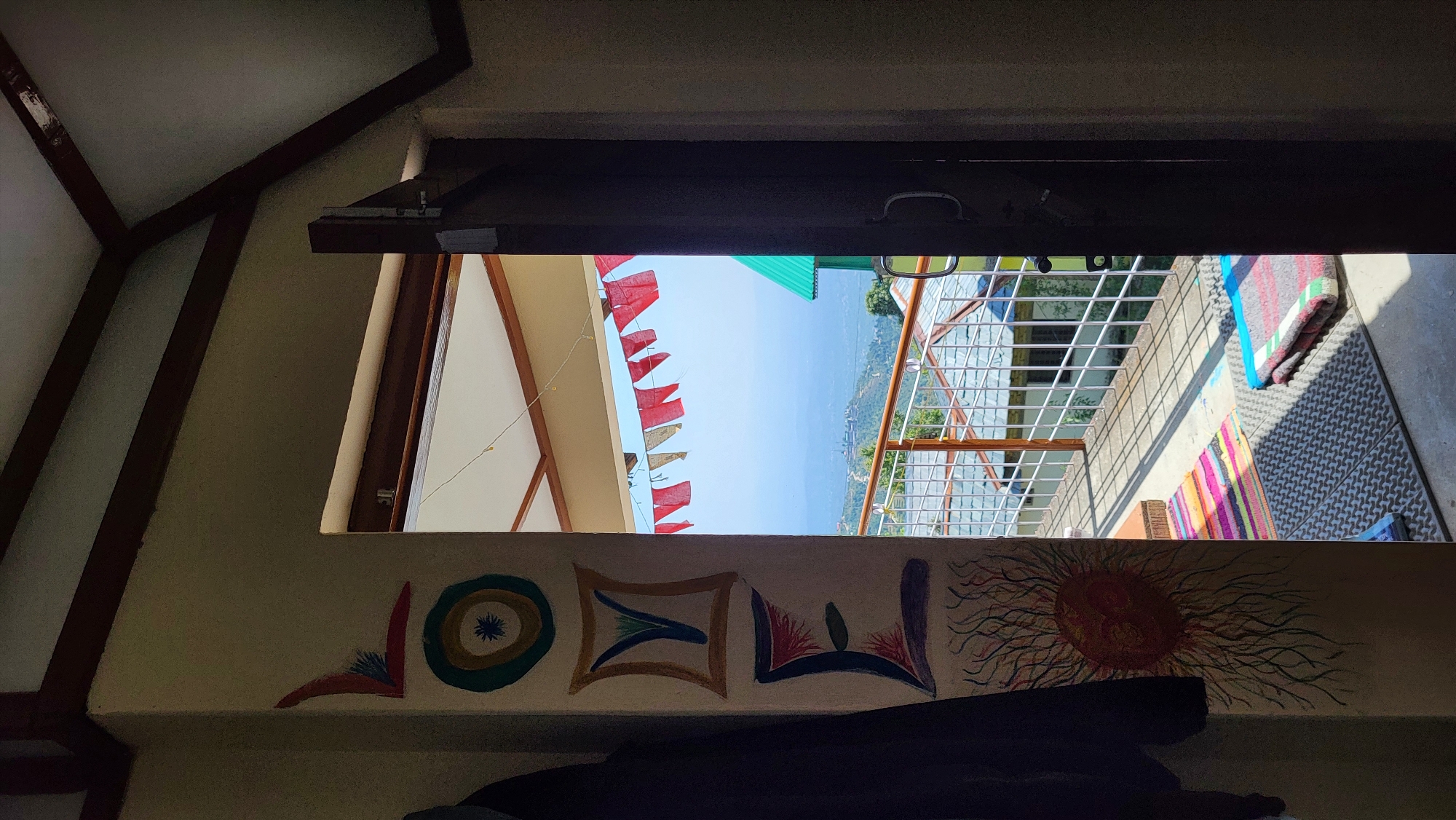 The photograph shows a view from inside a building, looking outward through a window. The window is partially open, revealing a landscape outside. Red flags are strung up on a building outside the window. Beyond the building is a mountain range and blue sky. The interior of the building is visible on the left side of the frame, and has a sloping roof. A white wall below the window is decorated with a series of colorful, stylized drawings, including an eye design, a rectangular shape, a bird and a sun. The location is likely Haini, India, based on the prompt. There are blankets and rugs visible on an outside patio.