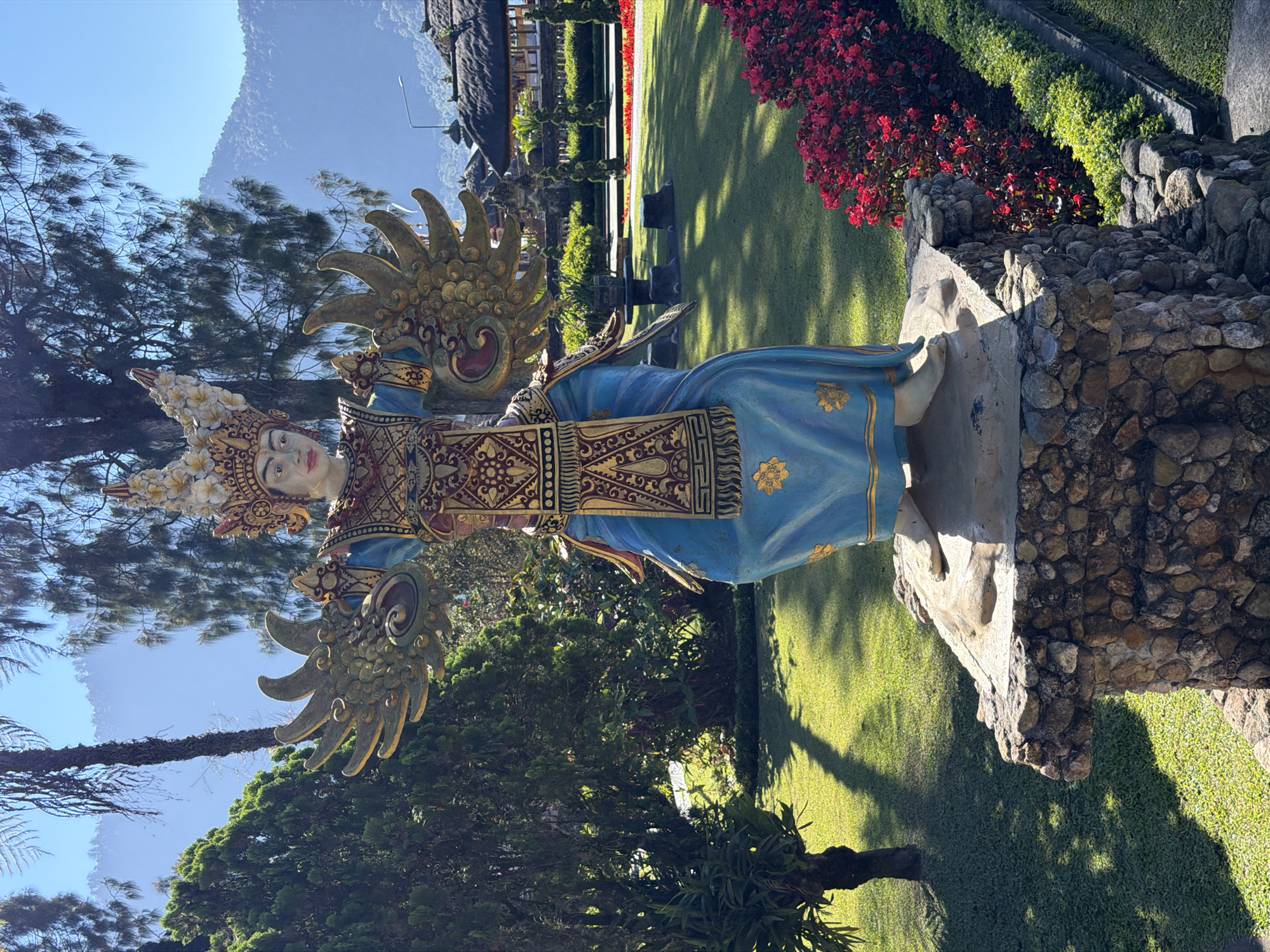 A full-shot, eye-level outdoor photograph captures a colorful statue of a mythical figure in a garden setting. The statue, predominantly blue with gold accents, stands on a stone pedestal. It is adorned with an elaborate headdress and gold wings. The figure is depicted with arms outstretched, hands in a gesture of welcome or blessing. The background shows lush green grass, manicured hedges, red flowers, trees, and a hazy mountain range under a clear blue sky. In the distance, traditional Balinese-style buildings with thatched roofs are visible. The scene suggests a tourist attraction or a cultural site in Baturiti, Indonesia. The lighting indicates a sunny day, casting shadows on the grass. There are no people in the immediate vicinity of the statue.