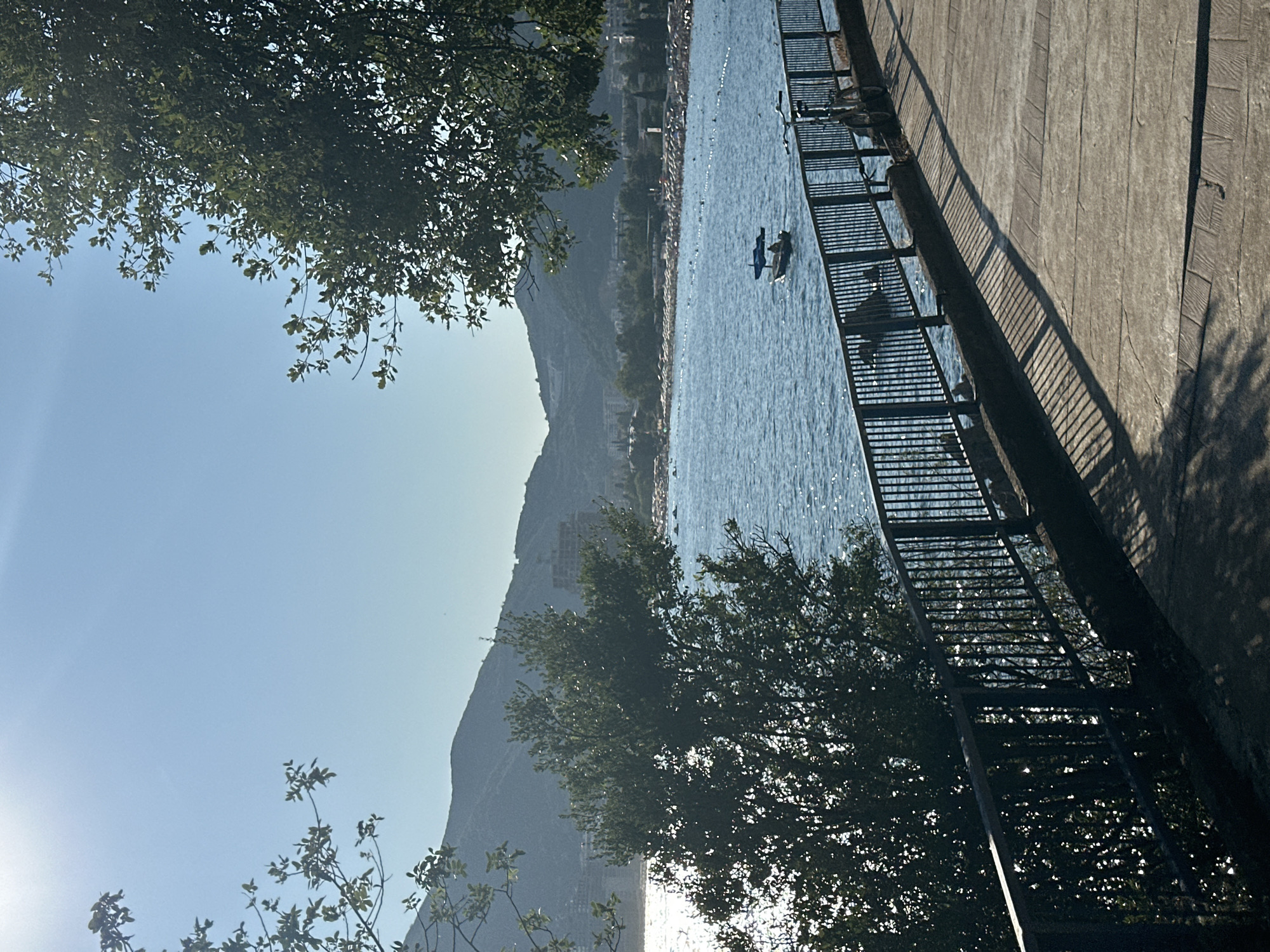 The image depicts a sun-drenched scene along the coast of Budva, Montenegro. A paved walkway runs parallel to a black metal railing, casting long shadows from nearby trees onto the path. Beyond the railing, the bright blue Adriatic Sea stretches out, reflecting the sunlight. A small boat with a blue canopy is visible on the water, carrying at least one person. In the distance, a bustling beach is lined with numerous beach chairs and umbrellas, with many people visible enjoying the sunny weather. Behind the beach, a range of green, mountainous hills forms the backdrop of the city, with buildings and some higher-rise structures integrated into the slopes. The sky is a clear, vibrant blue, with a bright sun illuminating the scene, creating a shimmering effect on the water. The overall atmosphere is one of a busy, pleasant summer day at the seaside.