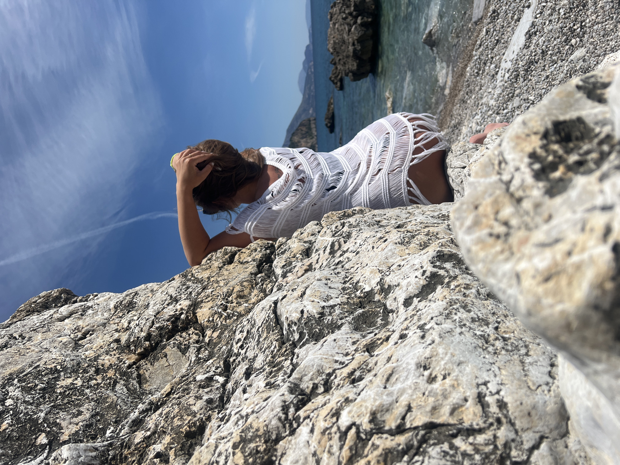 A woman is lying on a large rock, possibly near the Adriatic Sea. She wears a white crocheted cover-up. She is touching her hair with her left hand. The background includes a beach of small pebbles and the Adriatic Sea with distant mountains or hills. The location is Prijevor, Montenegro, as specified.