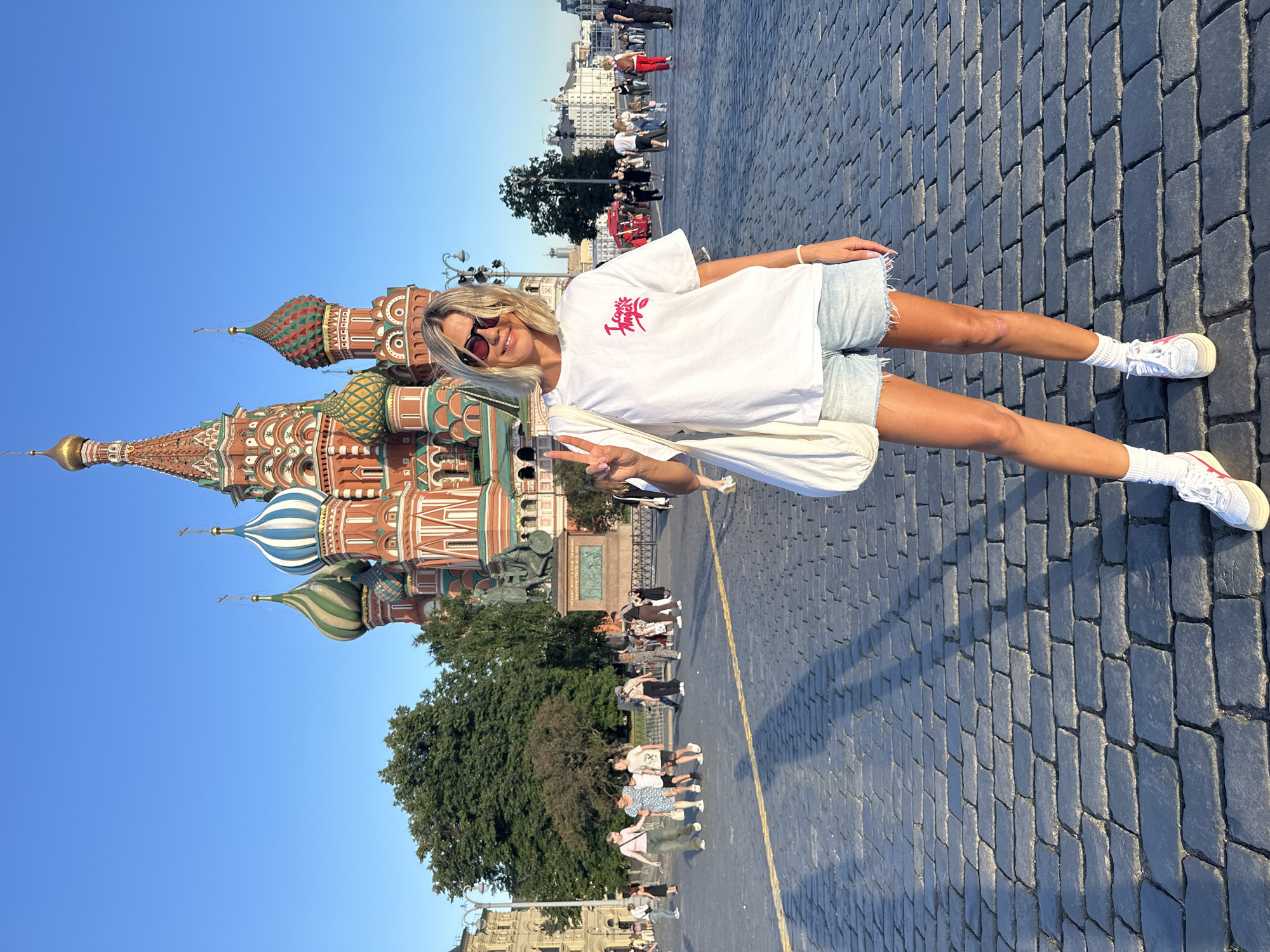 A woman with blonde hair and sunglasses stands in Red Square, Moscow, in front of Saint Basil's Cathedral. She is wearing a white t-shirt, denim shorts, white socks, and white sneakers. She is holding a white tote bag over her left shoulder and making a peace sign with her right hand. The cathedral's iconic colorful domes are visible behind her, as are other tourists and buildings in the background. The ground is paved with cobblestones, and the sky is clear and blue.
