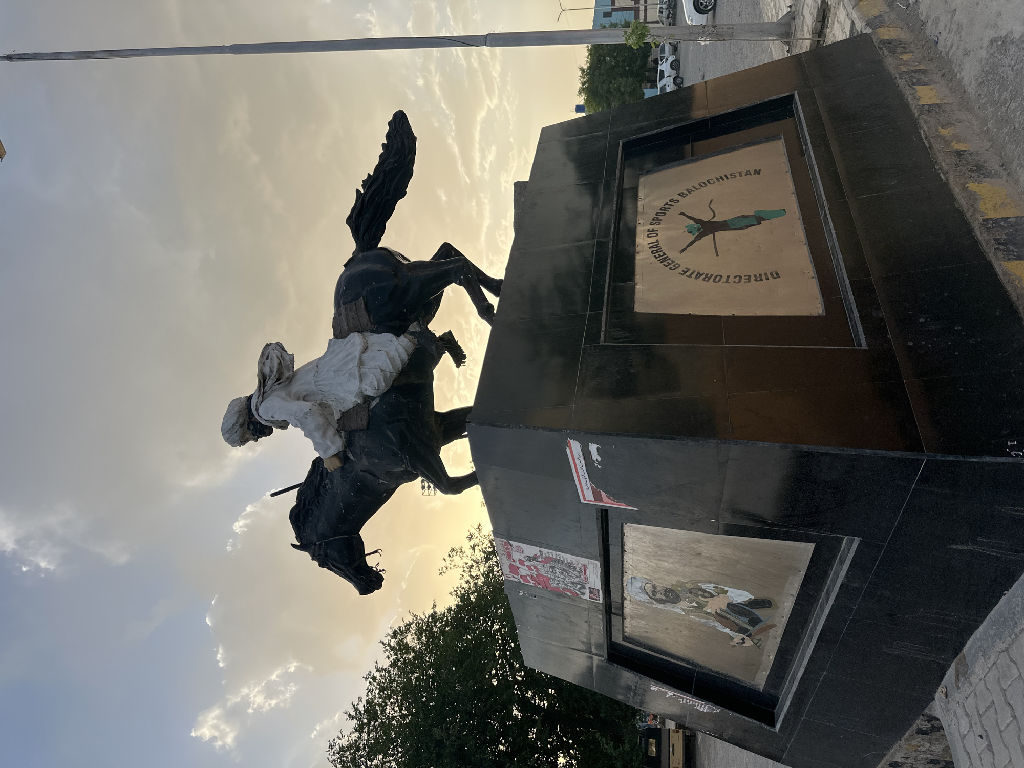 The image shows a statue of a man on a horse in Quetta City Tehsil, Pakistan. The statue is mounted on a black rectangular base. The base has three plaques on it, one of which reads "Directorate General of Sports Balochistan." The sky is cloudy with a hint of sunlight. There is a tree in the foreground. There are also cars and a building visible in the background. The statue is located on a paved area with a curb.