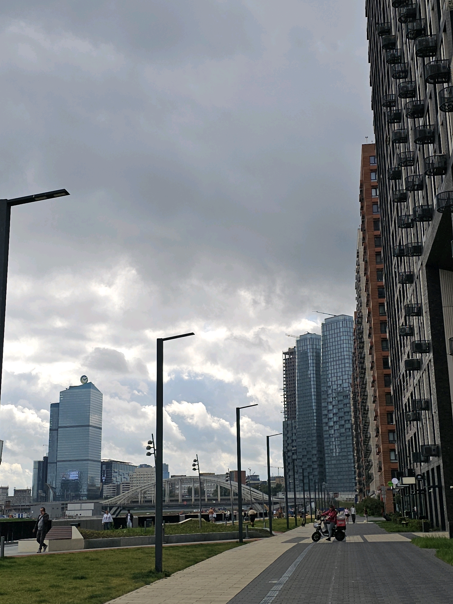 The image captures a view of the Moscow City business district under a cloudy sky. In the foreground, a paved walkway runs alongside a grassy area. Several modern streetlights line the path.
To the right, a tall, modern residential building with many balconies is prominently featured. Its facade is a mix of dark grey and reddish-brown tones. Further into the background, a cluster of other skyscrapers rises, with the distinctive blue glass building, likely the "Neva Towers," being a focal point. One of these buildings has a clock face at its peak.
Several people are visible in the scene. A man in a dark jacket is walking on the walkway near a bench. Further down the path, a person on a scooter, possibly a delivery person, is riding towards the viewer. Other individuals are seen walking in the middle ground, near the skyscrapers and a bridge.
The overall atmosphere is urban and contemporary, with a focus on modern architecture and infrastructure within a bustling city environment. The cloudy sky suggests a diffused daylight.