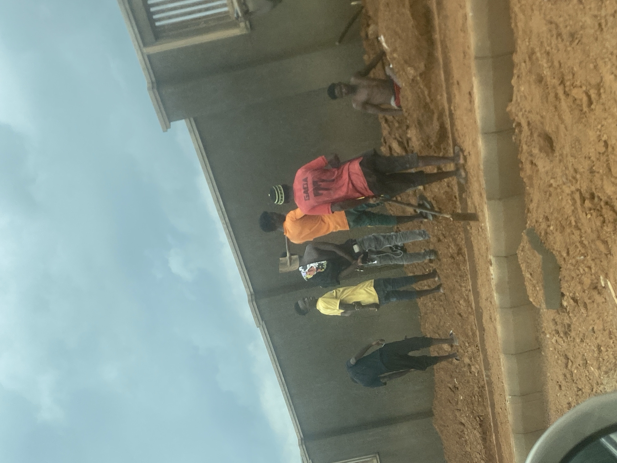 The image captures a group of approximately six men gathered on a dirt-covered worksite next to a concrete building, under an overcast sky. The photograph is taken from a slightly elevated perspective, looking down at the subjects and the ground.
The scene depicts an outdoor setting, likely during daytime, characterized by a predominantly brown, uneven dirt ground. A light-colored concrete curb or pathway runs vertically through the middle-right of the frame, separating different sections of the ground.
The men are spaced along the edge of the building on the left. From right to left: one man is shirtless, wearing red shorts, standing on a higher earthen bank. Next to him, another man wears a red t-shirt with the word "GADA" printed in white on its back, possibly holding a tool that extends downwards. Following him is a man in an orange t-shirt, and then another in a dark hoodie with a colorful graphic. A fifth man is seen in a bright yellow t-shirt and dark trousers, facing towards the left. The man furthest to the left is dressed in a dark t-shirt and dark trousers, with his back partially turned.
The building is a concrete structure with visible architectural details like horizontal slats on its upper levels. The sky is a pale, cloudy blue, suggesting an overcast day. The overall impression is one of laborers or workers at a construction or development site in Gosa, Nigeria.