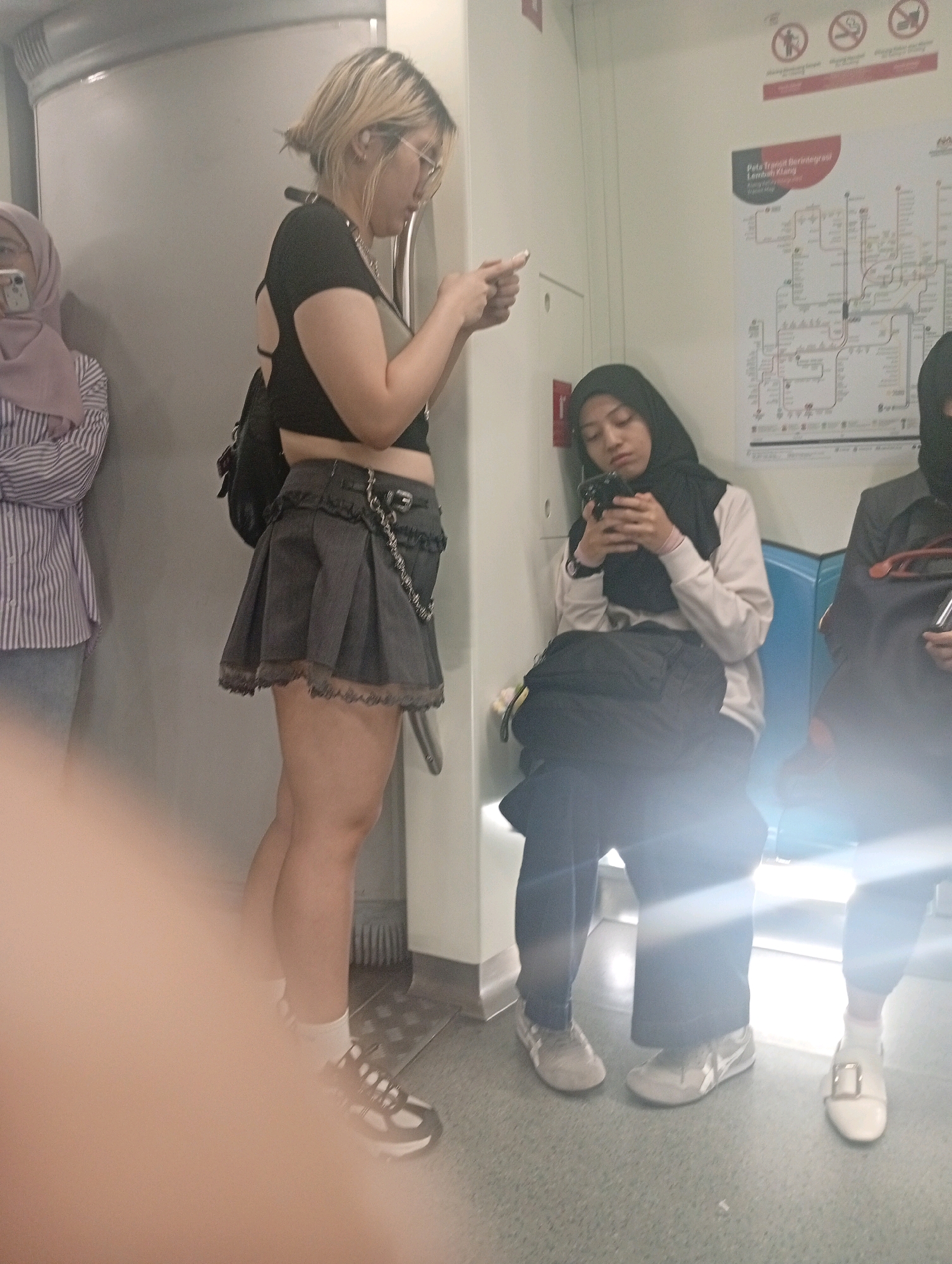 An interior shot taken from a low angle on what appears to be public transportation, possibly a train. The lighting is bright and there are lens flares. On the left, a woman with short blonde hair, wearing glasses and a black top with a cut-out back and a gray pleated skirt, is standing and looking at her phone. She has a backpack on her shoulders and is wearing white socks and black and white sneakers. To her right, a woman wearing a black hijab and a light-colored sweater is seated, also looking at her phone. She is wearing dark pants and white sneakers. Further to the right, a person wearing a black face mask and a dark jacket is partially visible, holding a red object. A map of the "Peta Transit Berintegrasi Lembah Klang" is displayed on the wall behind the seated woman. The floor is gray and speckled.