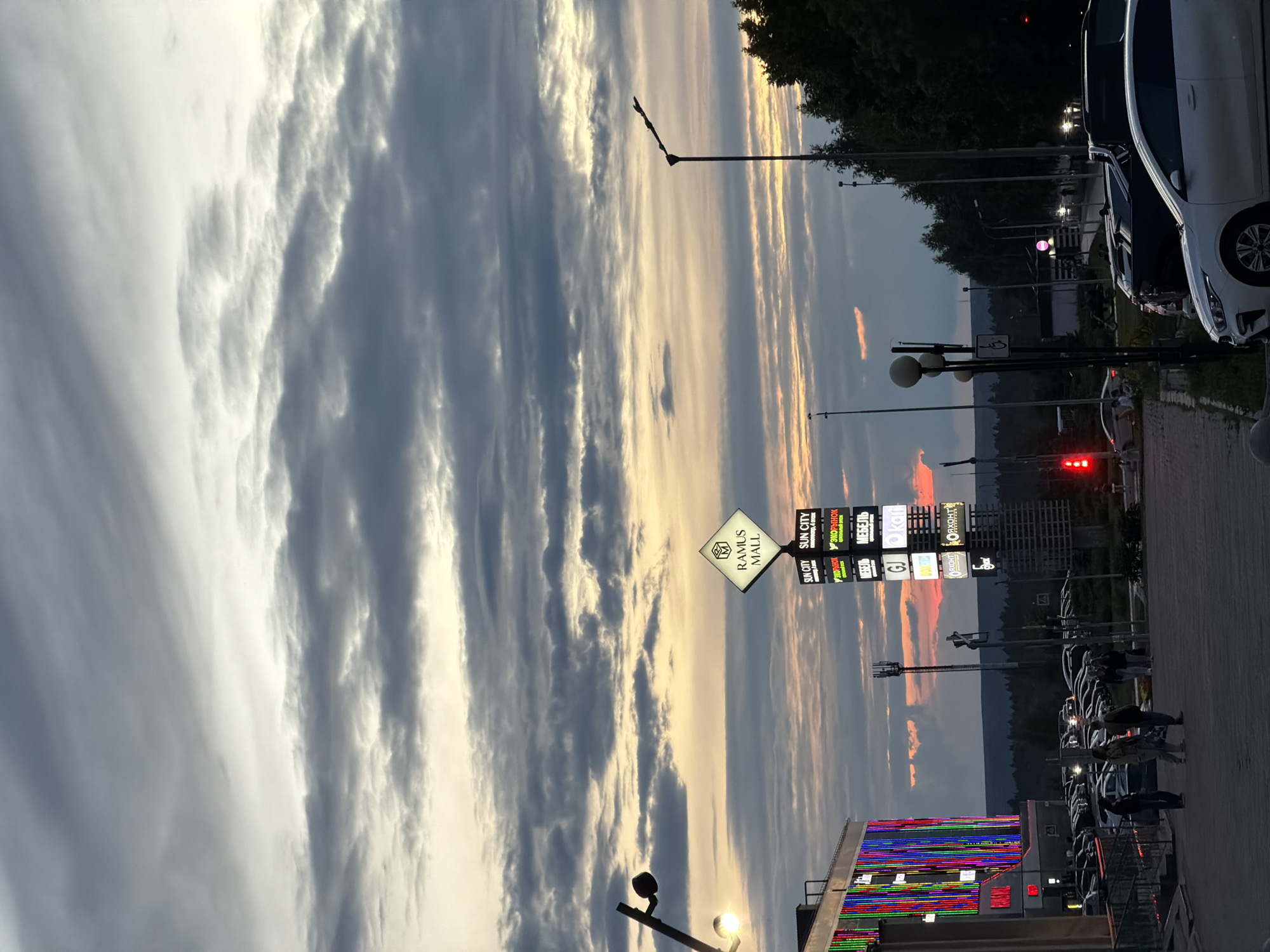The image depicts a street scene at dusk in Nizhnekamsk, Russia. The sky is filled with dramatic, layered clouds, illuminated by the setting sun, creating a warm glow. The foreground features a sidewalk and a parking lot with several cars. A prominent signpost displays the "RAMUS MALL" logo, and further down, a tall sign with multiple illuminated panels advertises various businesses, including "SUN CITY," "ЭКОРЫНОК" (Eco Market), and "МЕБЕЛЬ" (Furniture). Streetlights are visible, and one is illuminated. Several people are walking along the sidewalk, suggesting a leisurely evening. The overall atmosphere is calm and hints at the transition from day to night. The presence of the mall and various shops indicates a commercial and public space.